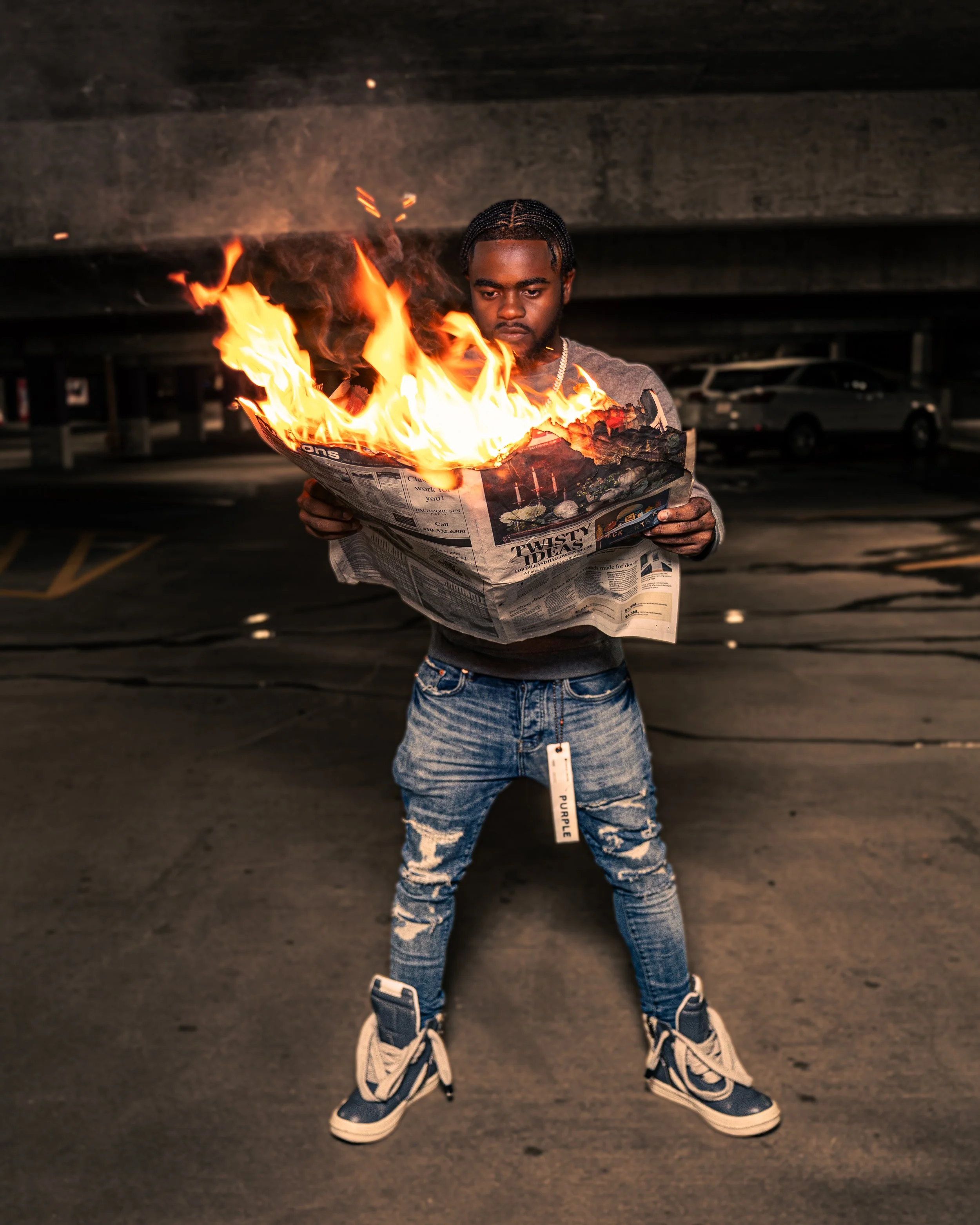 A young man with braided hair wearing ripped jeans and high-top sneakers stands in a parking garage, holding a burning newspaper.