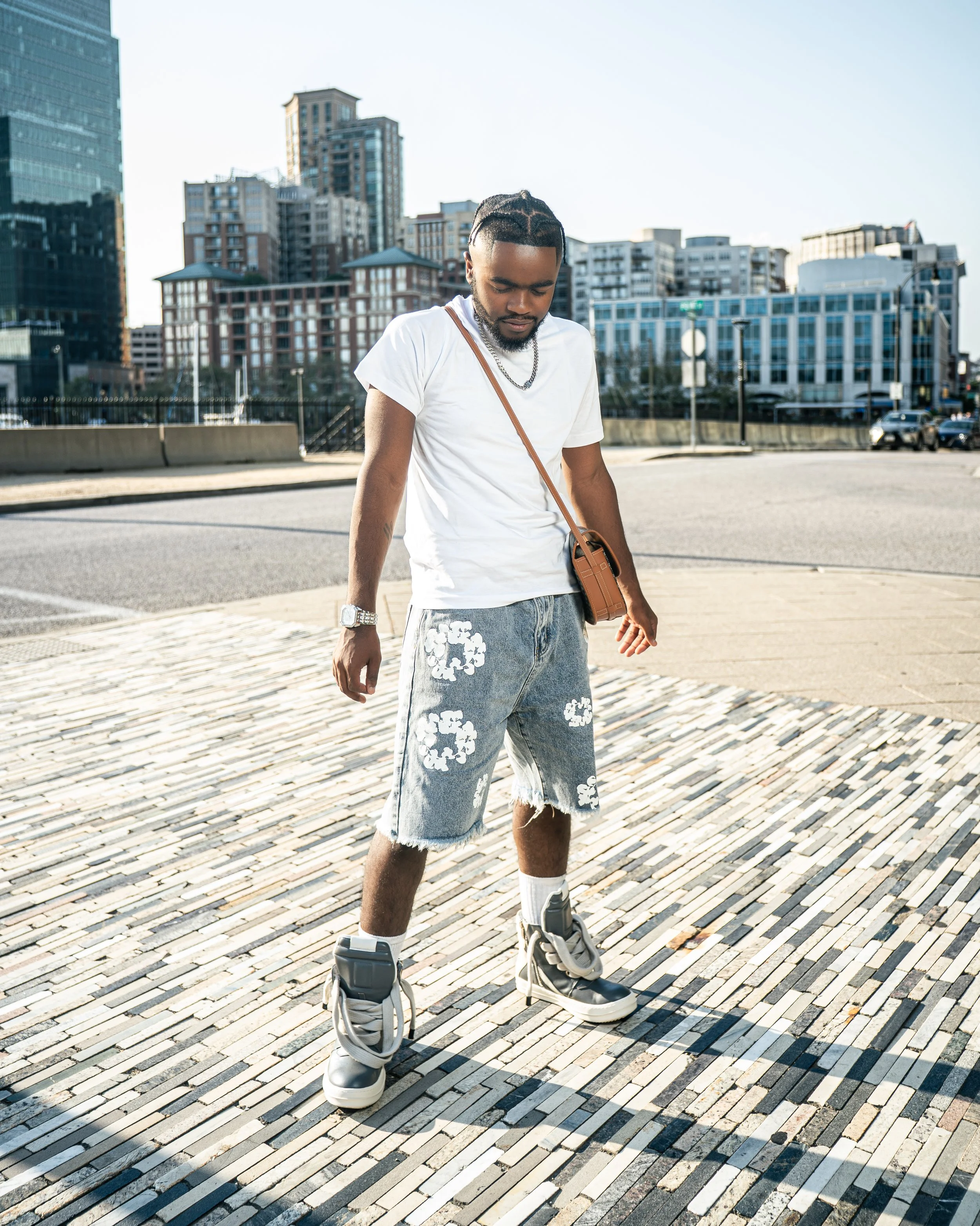 A man in a white t-shirt and denim shorts with cloud patterns stands on a paved area in an urban setting, wearing roller shoes, with tall buildings in the background.