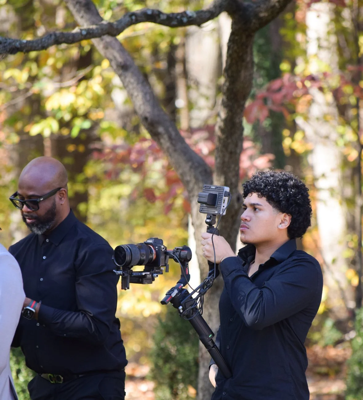 Two men filming outdoors in a wooded area during autumn. One man with curly hair and earrings holds a camera stabilizer, while the other man with glasses and a beard is focused on something off-camera.