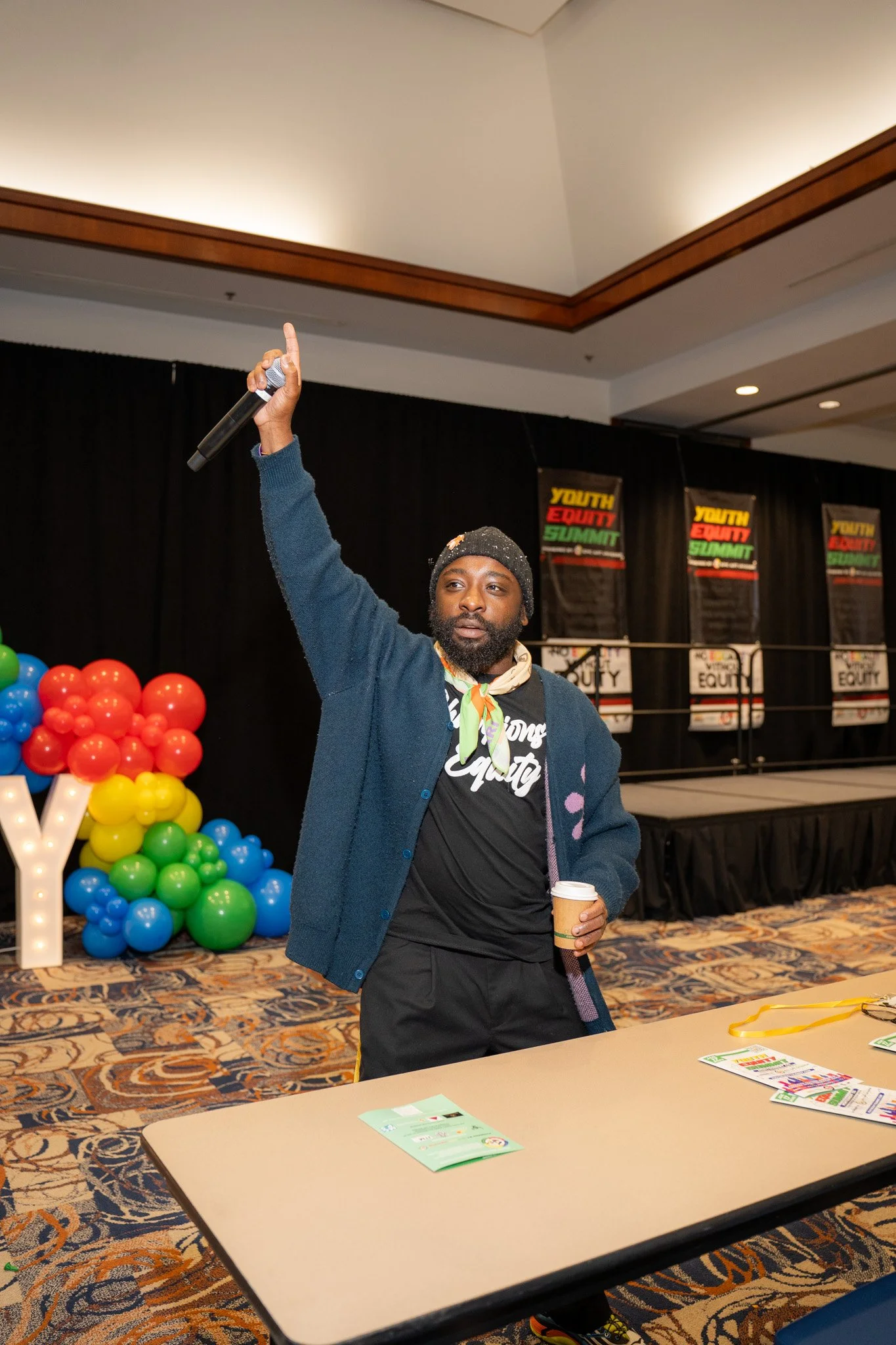 A man standing at a table in a conference room, holding a coffee cup in his left hand and raising his right hand with one finger pointing up. Behind him, there are colorful balloons and banners that read 'Youth Equity Summit', with a stage and carpet