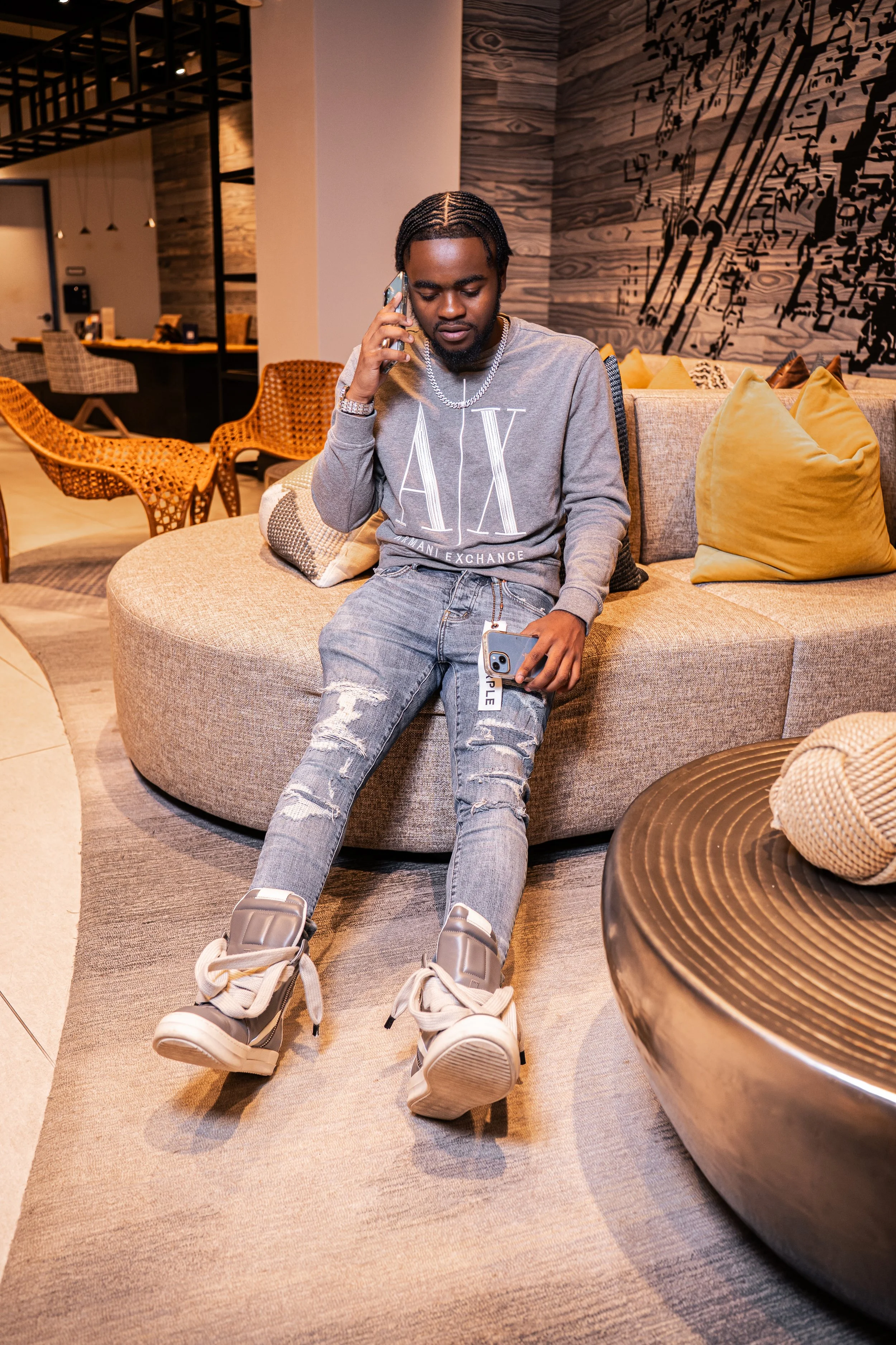 A young man sitting on a curved beige sofa in a modern, stylish lobby, speaking on a cell phone, holding another phone in his lap, wearing a gray Armani Exchange sweatshirt, ripped jeans, and sneakers.