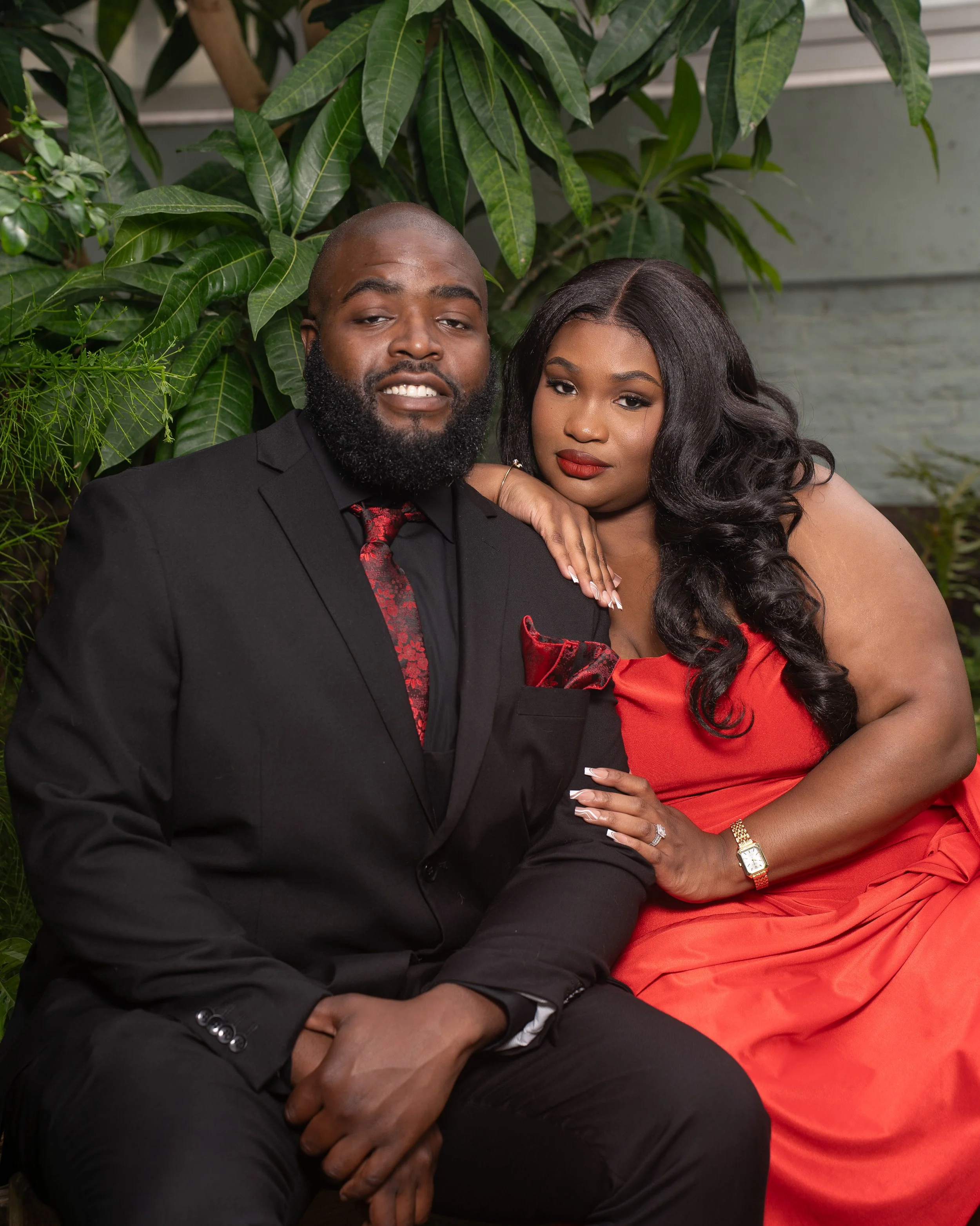A well-dressed couple posing together in front of green leafy plants. The man wears a black suit with a red patterned tie and pocket square, and the woman wears a strapless red dress with long, wavy hair, makeup, and jewelry.