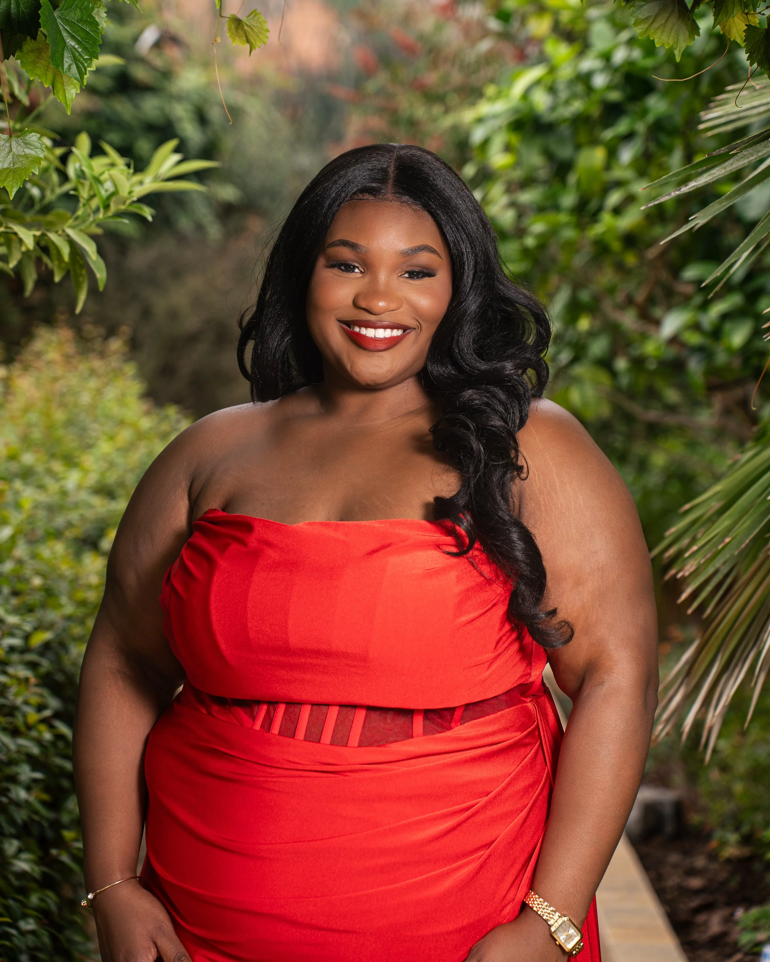 A smiling woman with long black hair wearing a red strapless dress standing outdoors among green plants and trees.