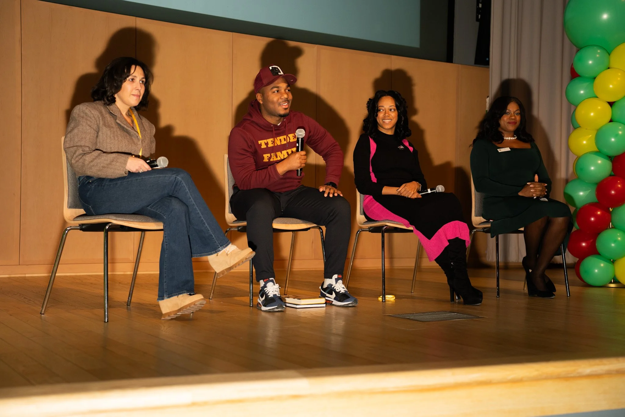 Four people sitting on chairs on a stage, participating in a panel discussion. The woman on the far left has dark hair, beige jacket, jeans, and beige shoes. The man second from left wears a maroon hoodie, black pants, and sneakers. The woman second 