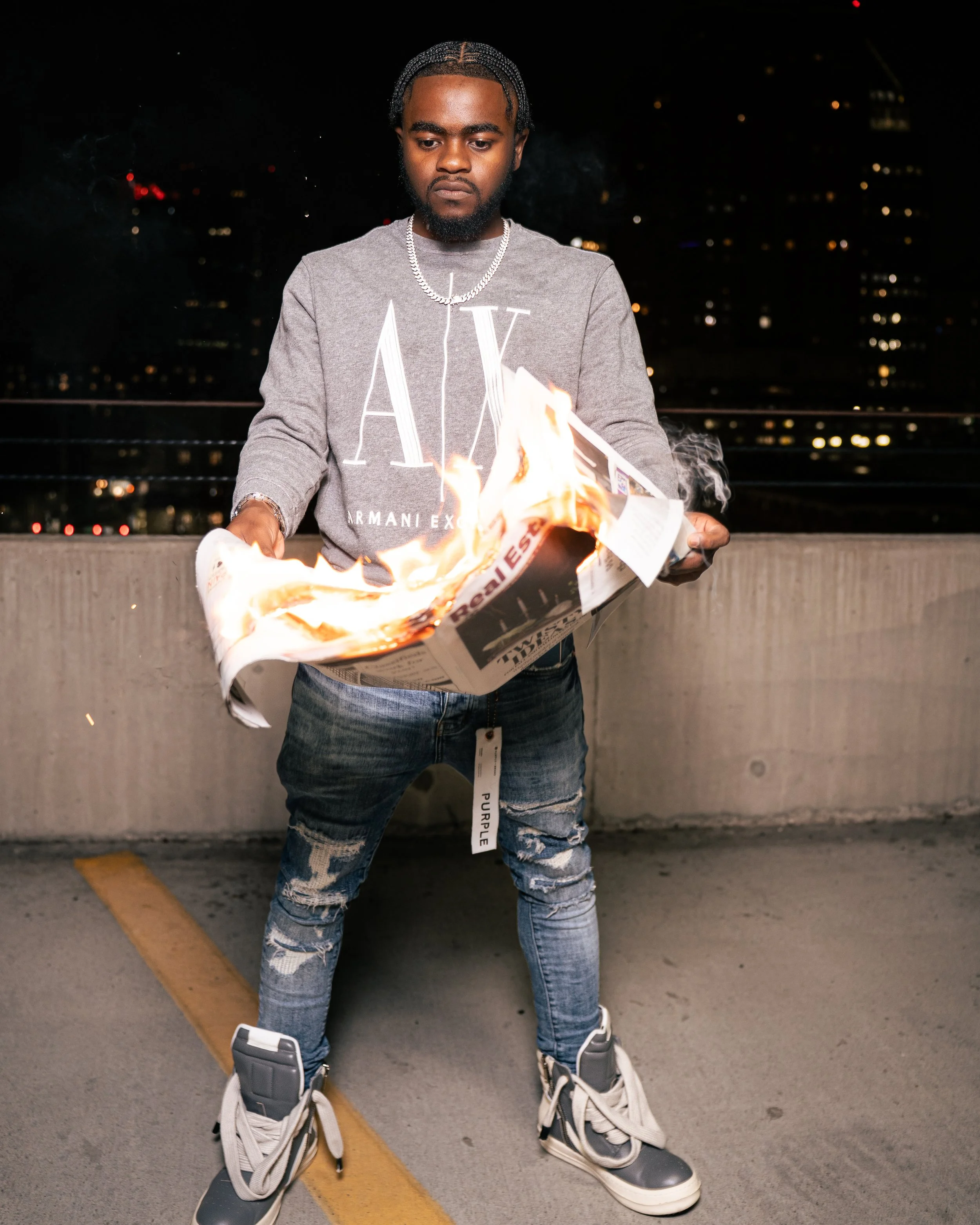 A young man with braided hair and a beard burns a paper newspaper on a rooftop parking lot at night.