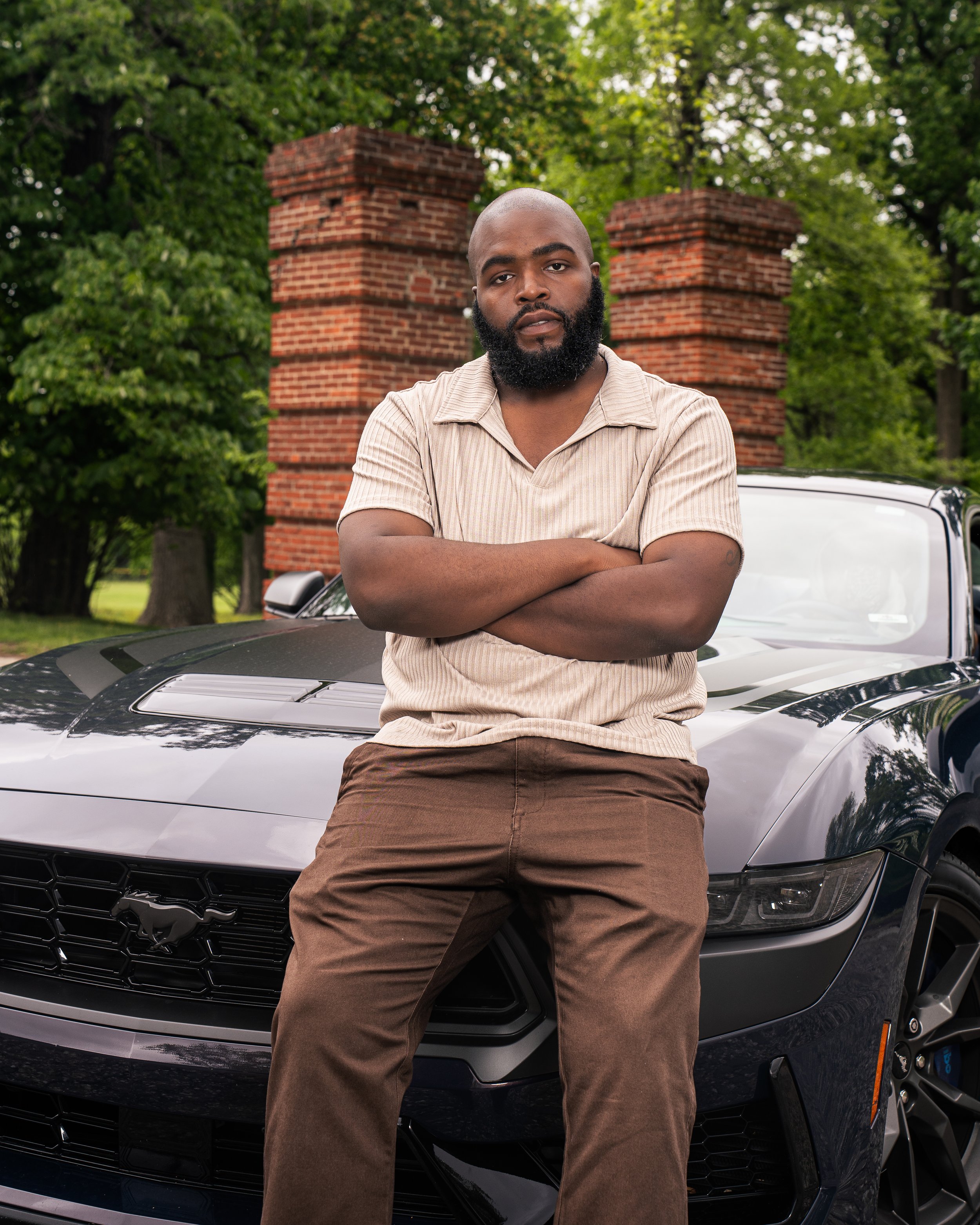 A man with a beard and bald head wearing a beige striped shirt and brown pants, standing with arms crossed in front of a black Ford Mustang parked outdoors. There are red brick columns and green trees in the background.