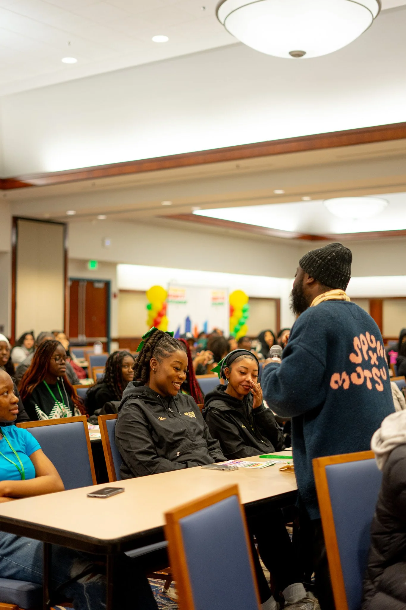 A man holding a microphone speaking to a group of young women seated at tables in a conference room, with many other people in the background.