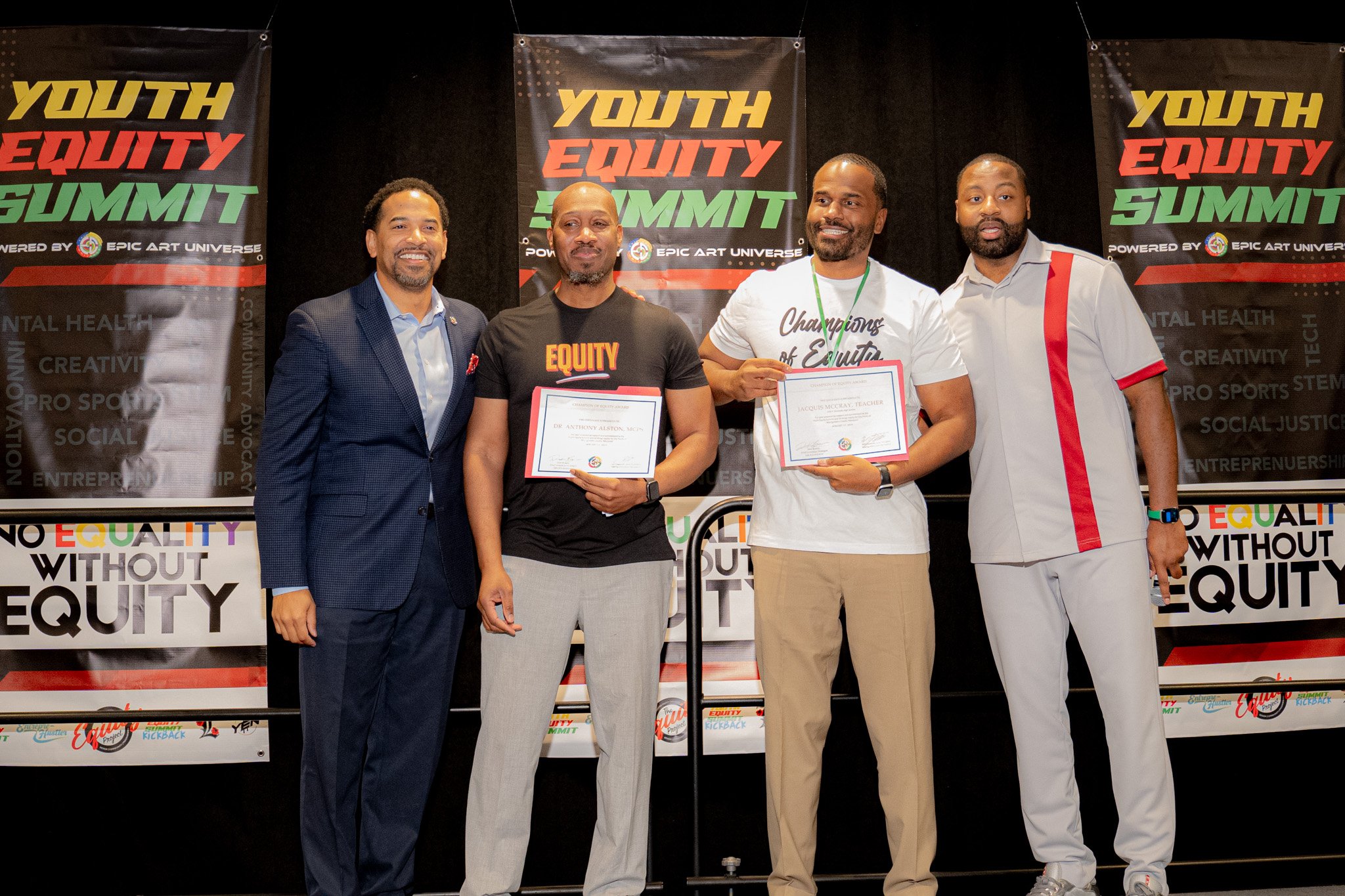 Four men standing on stage at the Youth Equity Summit, two of whom hold framed certificates. The background features banners with the summit's logo and topics like mental health, creativity, sports, social justice, and entrepreneurship.