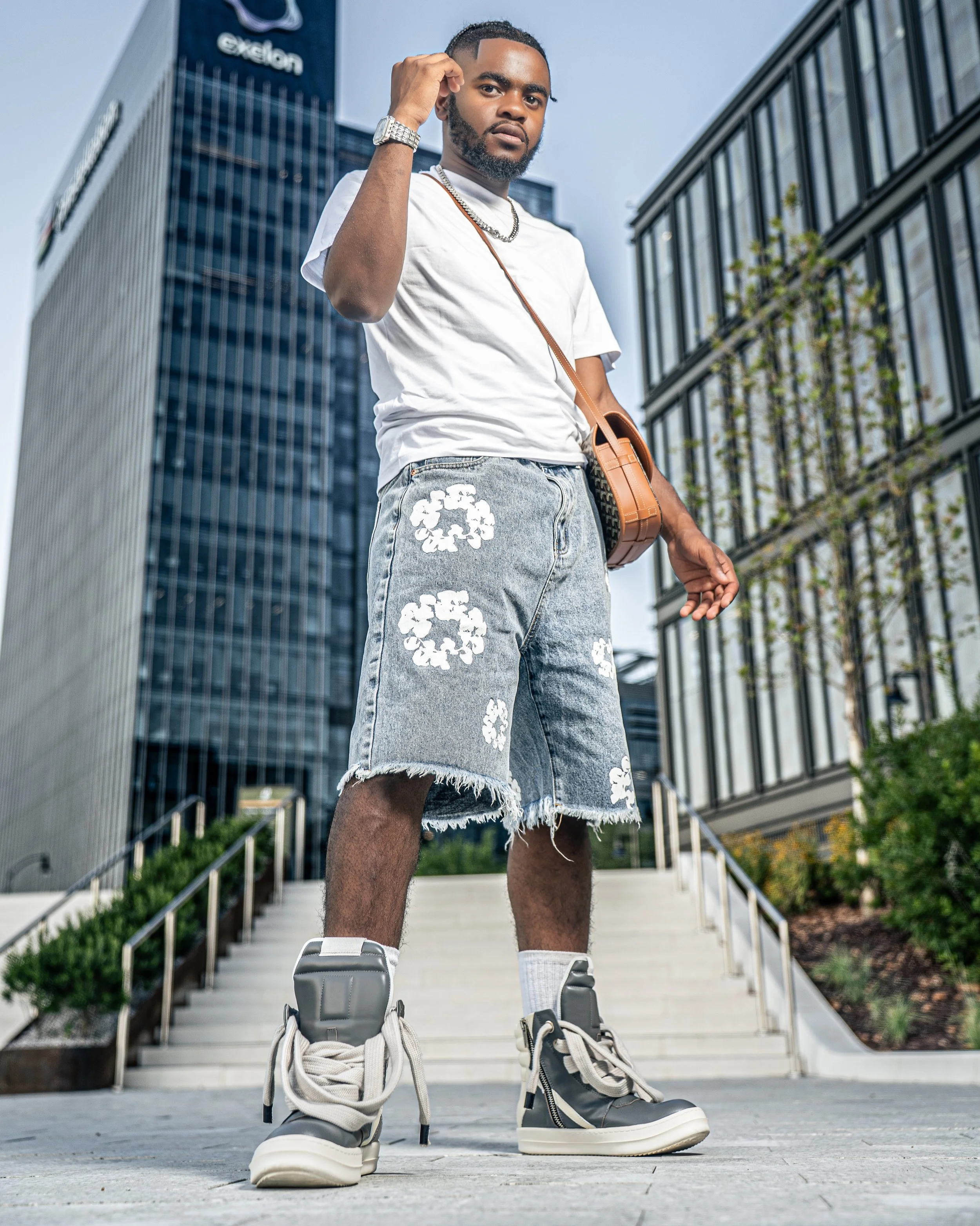 Young man standing outdoors in an urban setting with tall modern glass buildings, wearing a white t-shirt, frayed denim shorts with white floral patterns, high-top sneakers, and accessories including a watch, necklace, and purse.