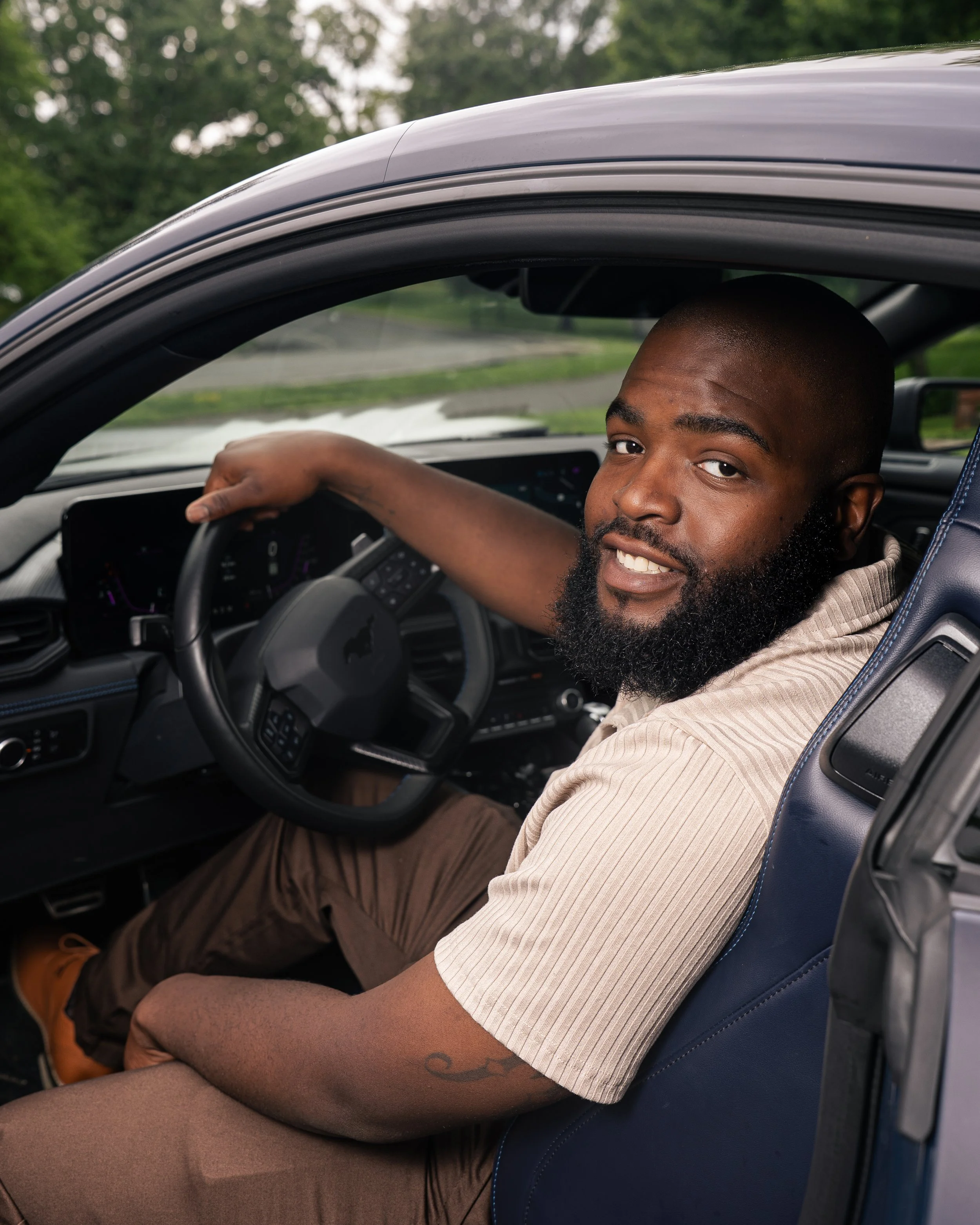 A man with a beard sitting in the driver's seat of a car, smiling at the camera, with his left arm resting on the steering wheel and the car window down.