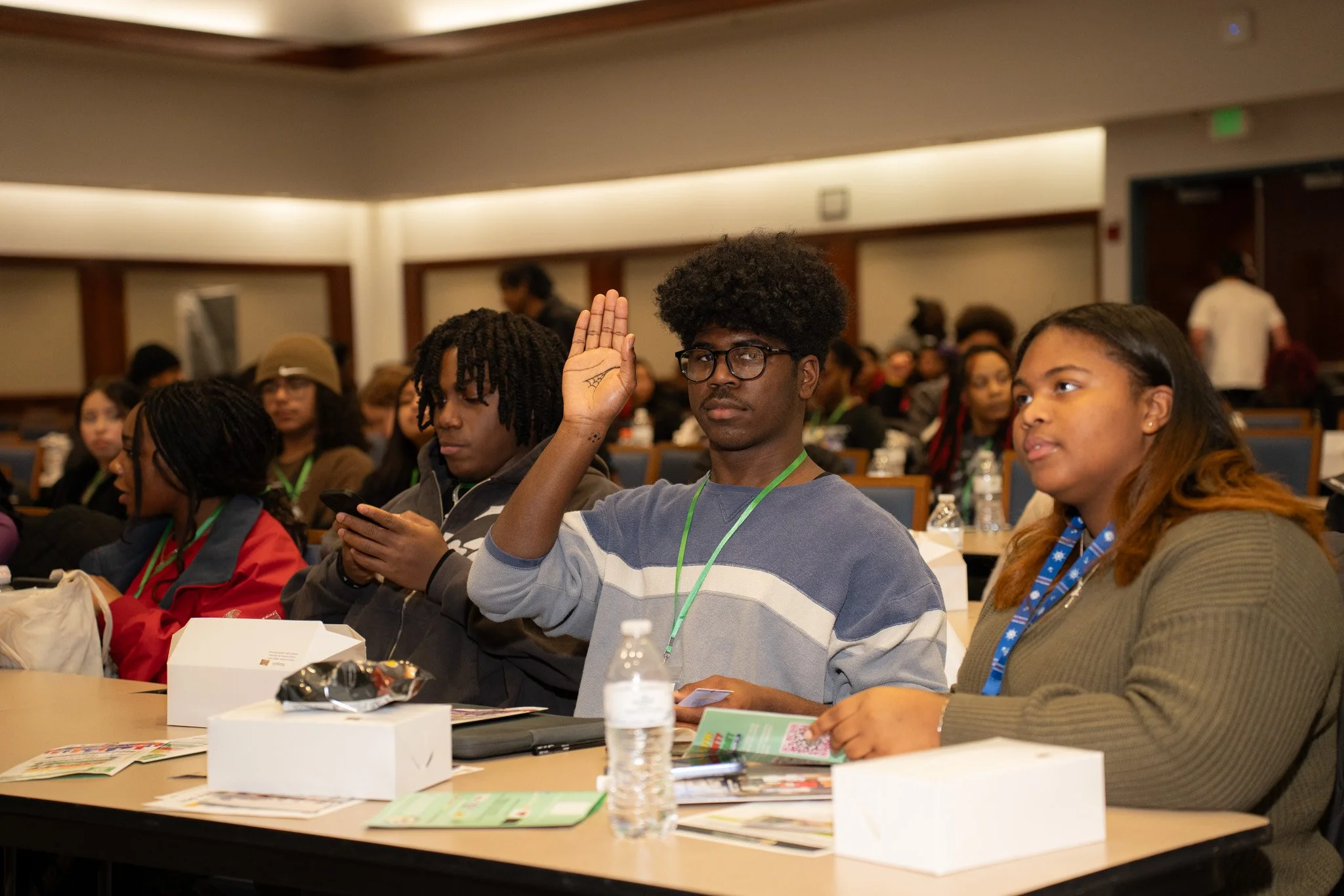 A young man with curly hair, glasses, and a gray and blue sweater raises his hand during a conference or seminar. Next to him, a young woman with long hair looks to the side. Other attendees are seated at the long tables, some using their phones. The