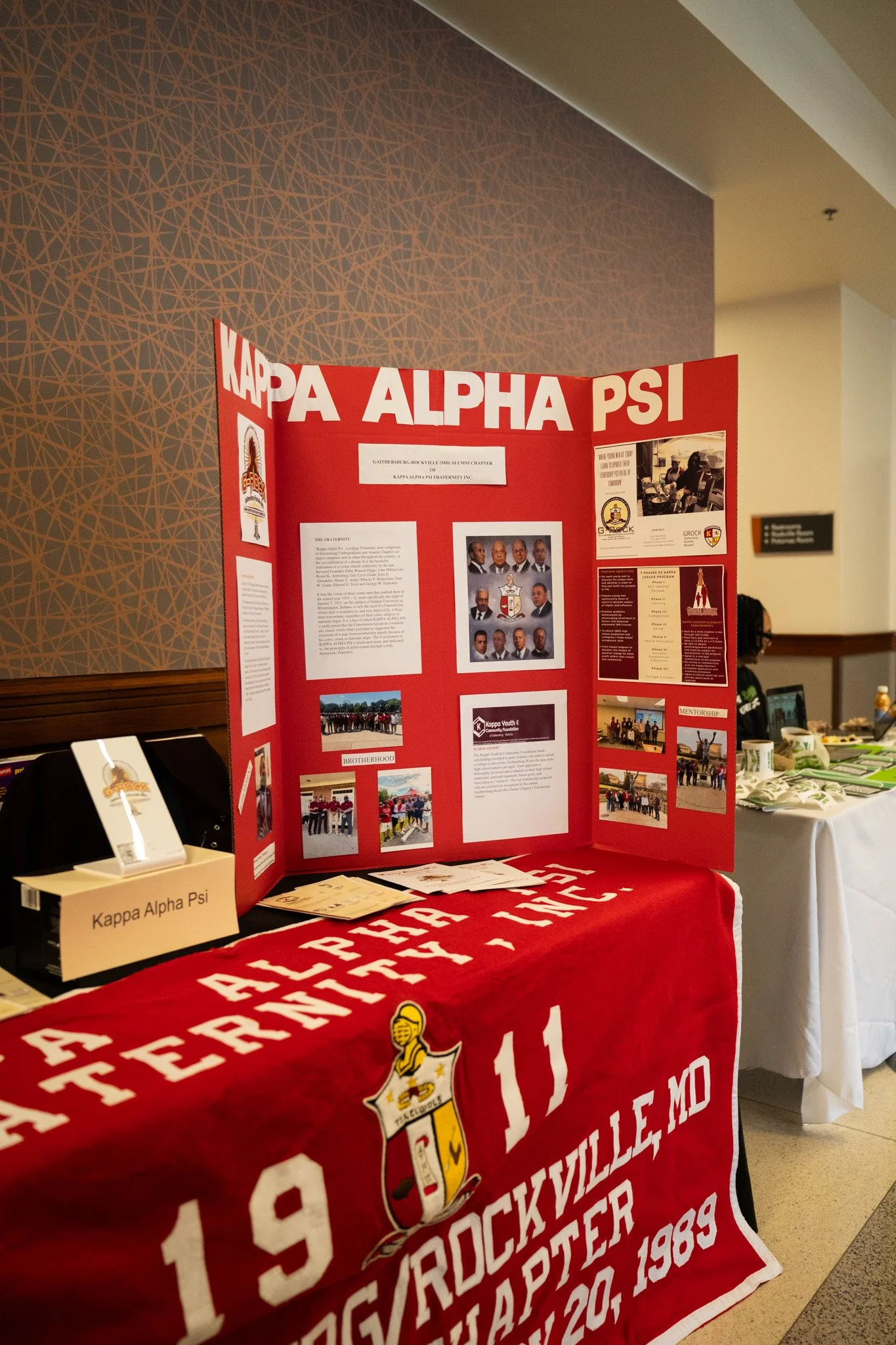 Display board and table with a red cloth, supporting Kappa Alpha Psi fraternity, Inc., information and photos at an event.