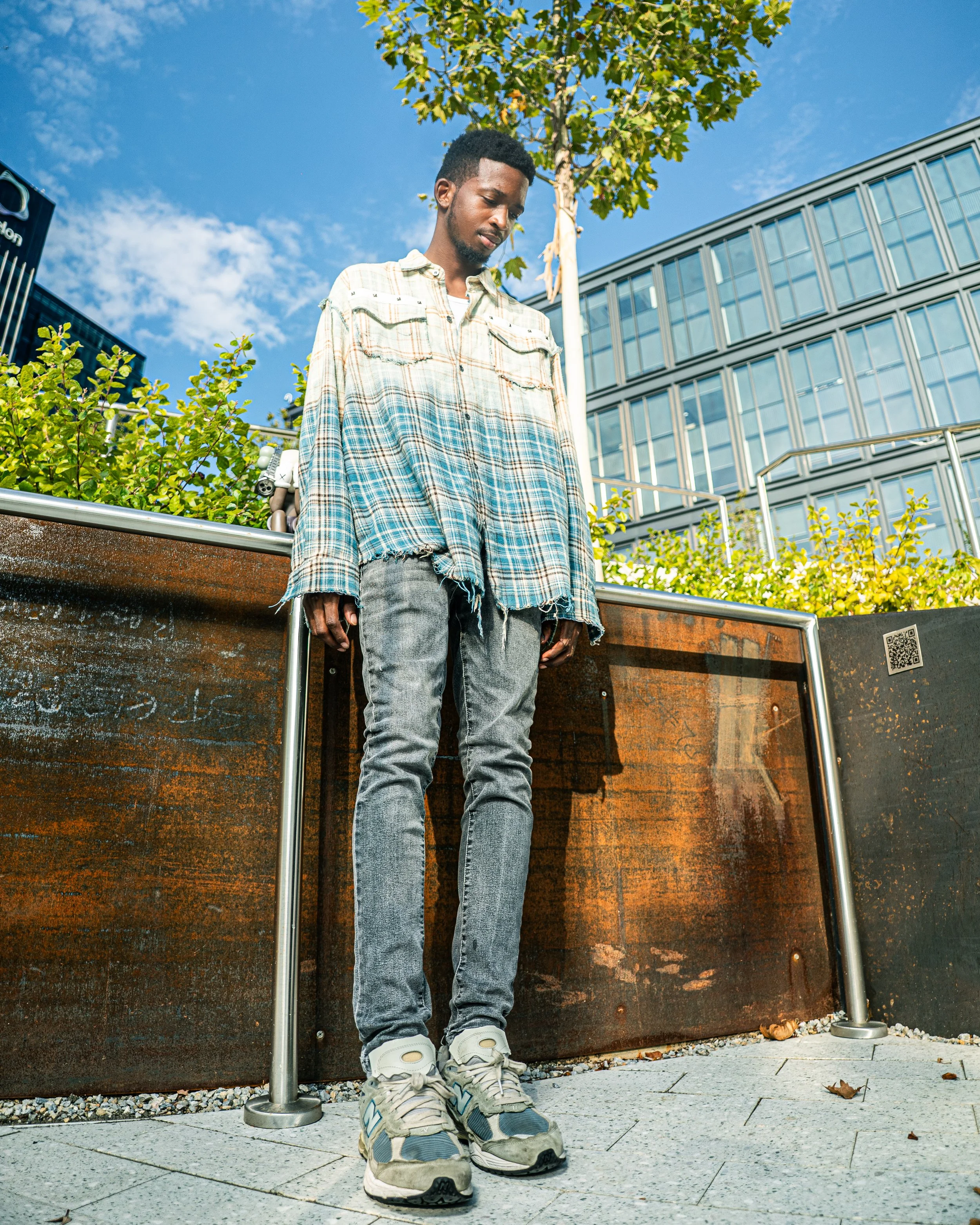 Young man standing outdoors in urban setting, wearing a plaid shirt, gray jeans, and sneakers, with modern buildings and a blue sky in the background.