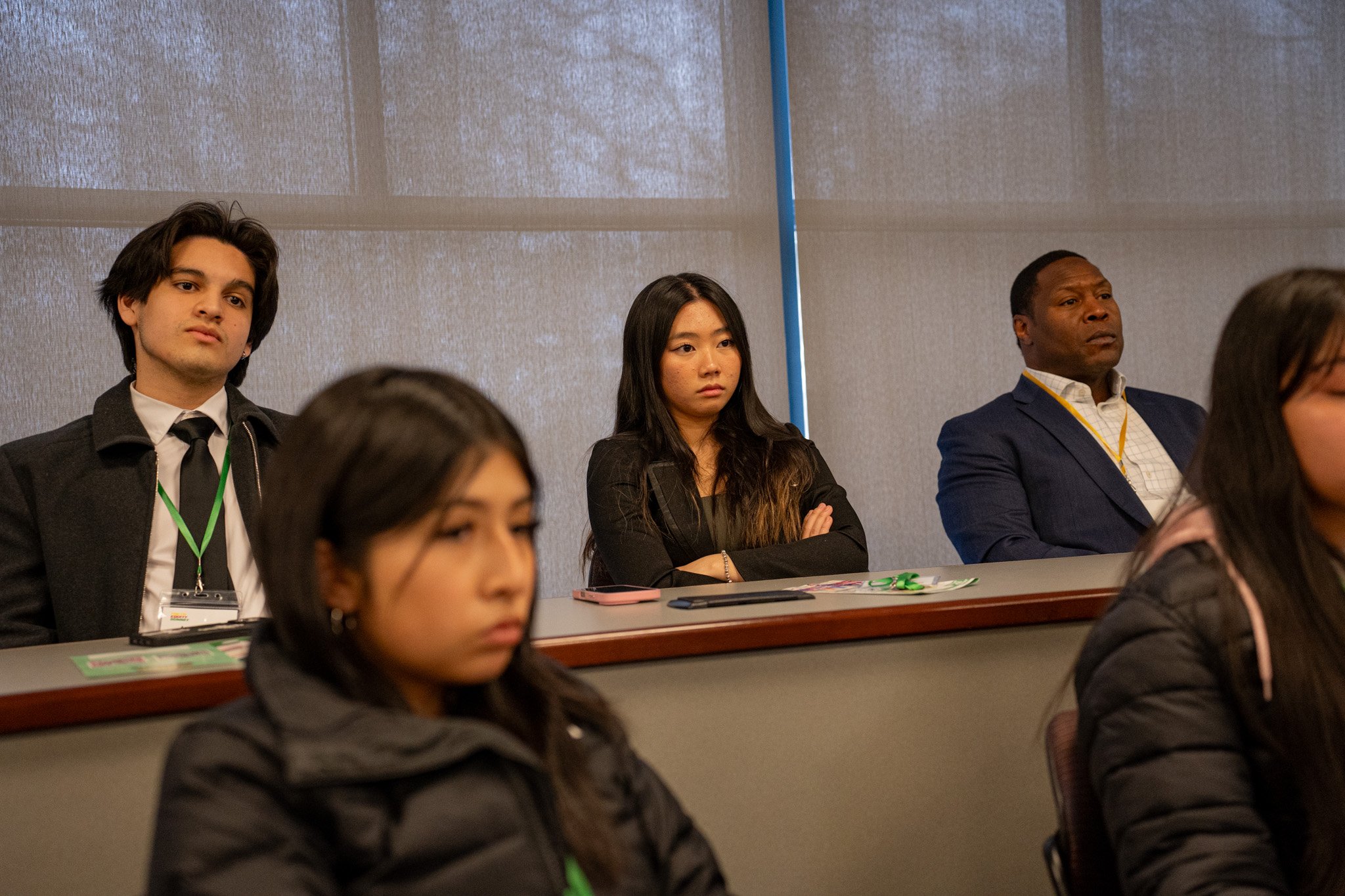 People attentively listening at a conference or seminar, seated behind a desk, with a plain background.