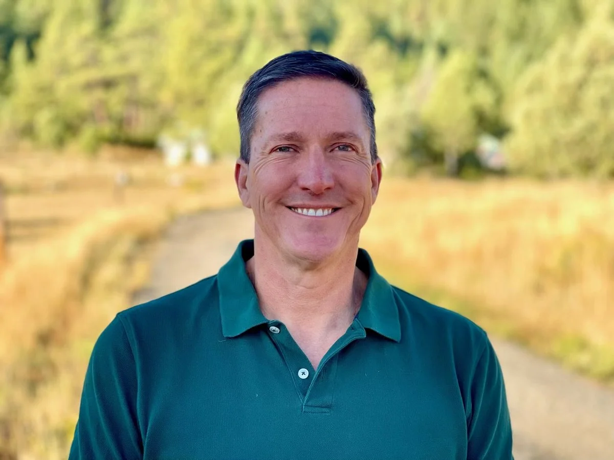 A smiling man with short dark hair wearing a green polo shirt outdoors with trees and a dirt path in the background.