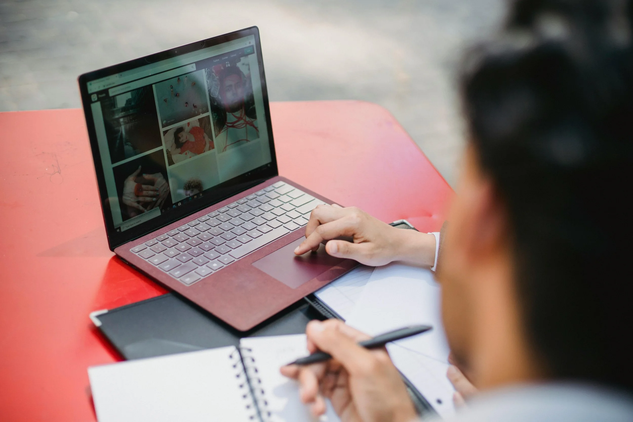 Person using a pink laptop with a backpack nearby, taking notes with a pen on paper while browsing social media or photo gallery on the laptop. Rethink to Rise AI