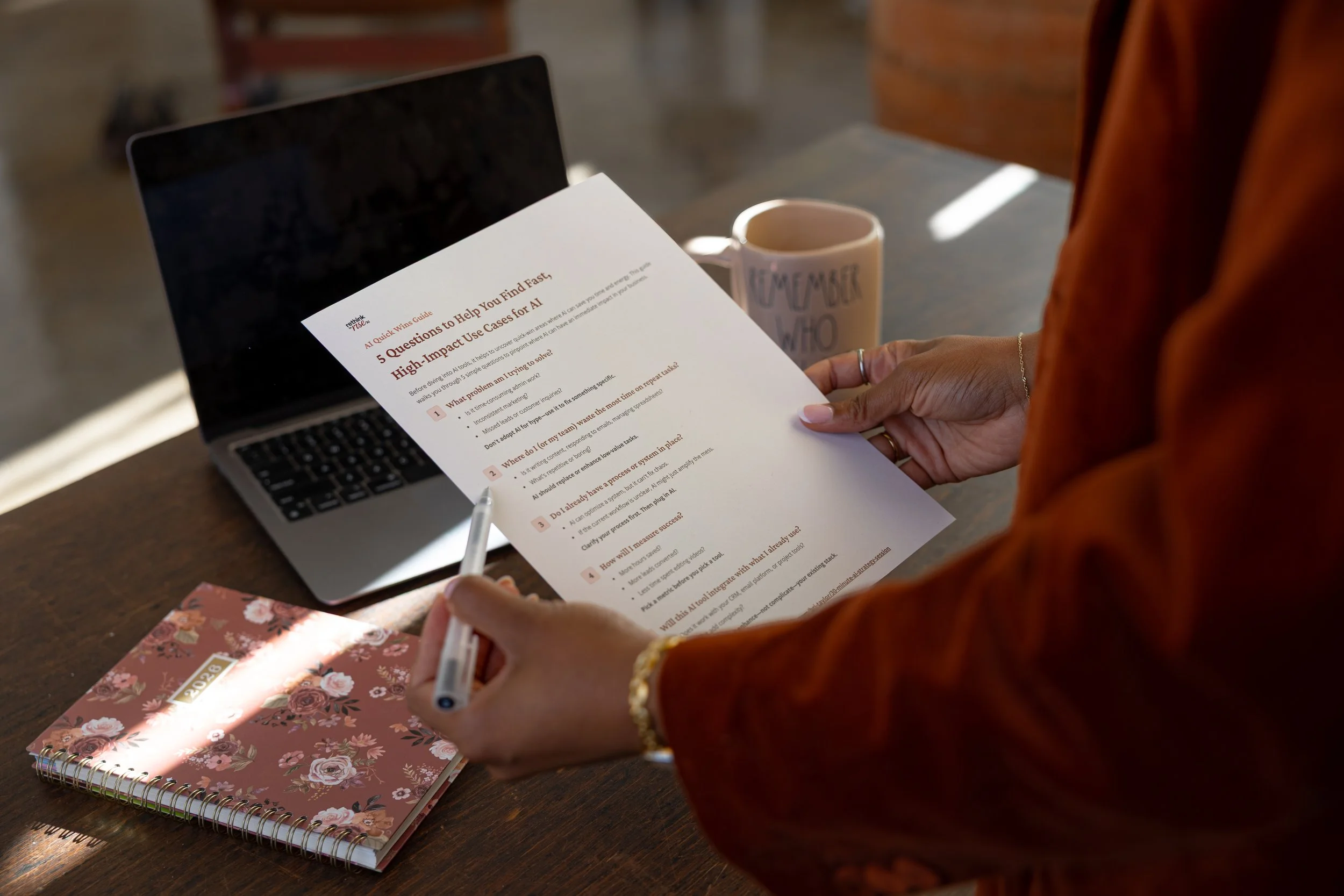 Person holding a printed list titled '5 Questions to Help You Find Fast, High-Impact Use Cases for AI' at a desk with a laptop, pink floral notebook, and a mug.