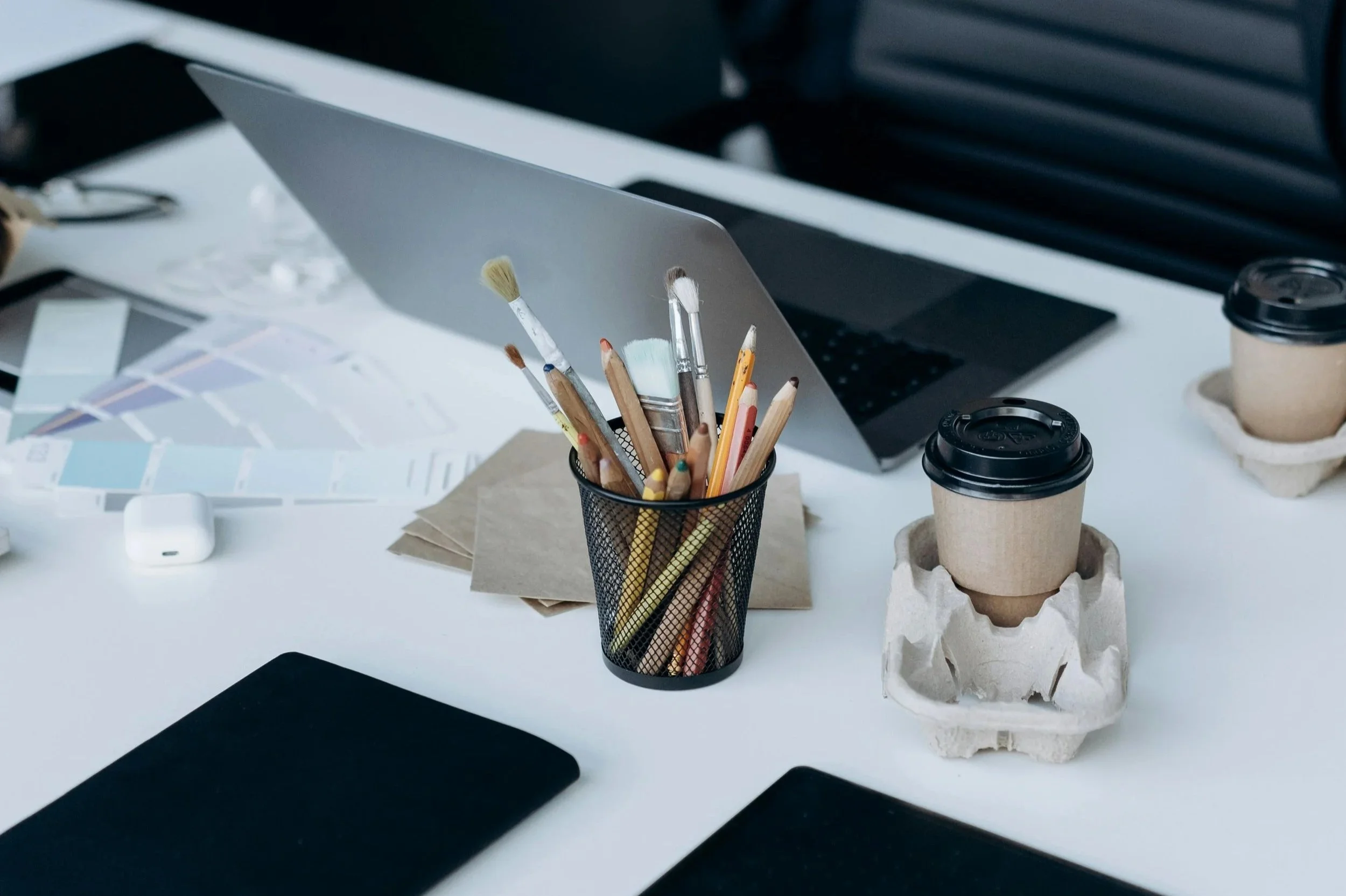 Desk with a laptop, paintbrushes in a black mesh holder, color swatches, paper envelopes, two to-go coffee cups in cardboard sleeves, and an AirPods case. Rethink to Rise AI