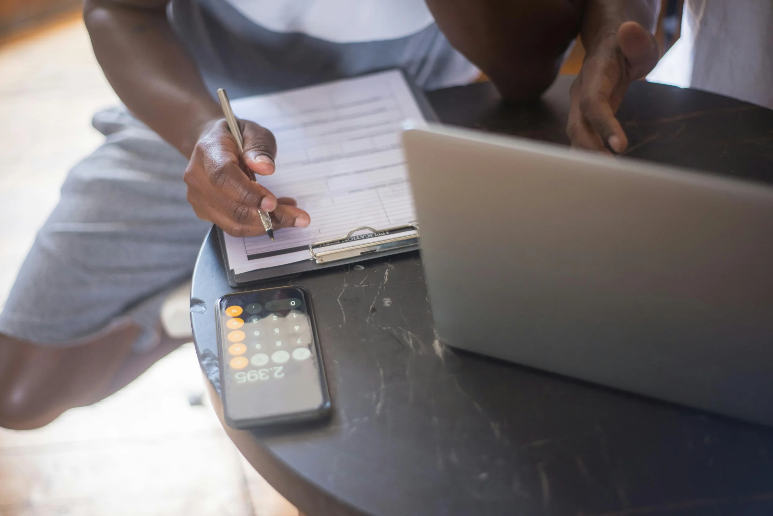 Person writing in a planner on a black table with a smartphone displaying numbers and a laptop. Rethink to Rise AI