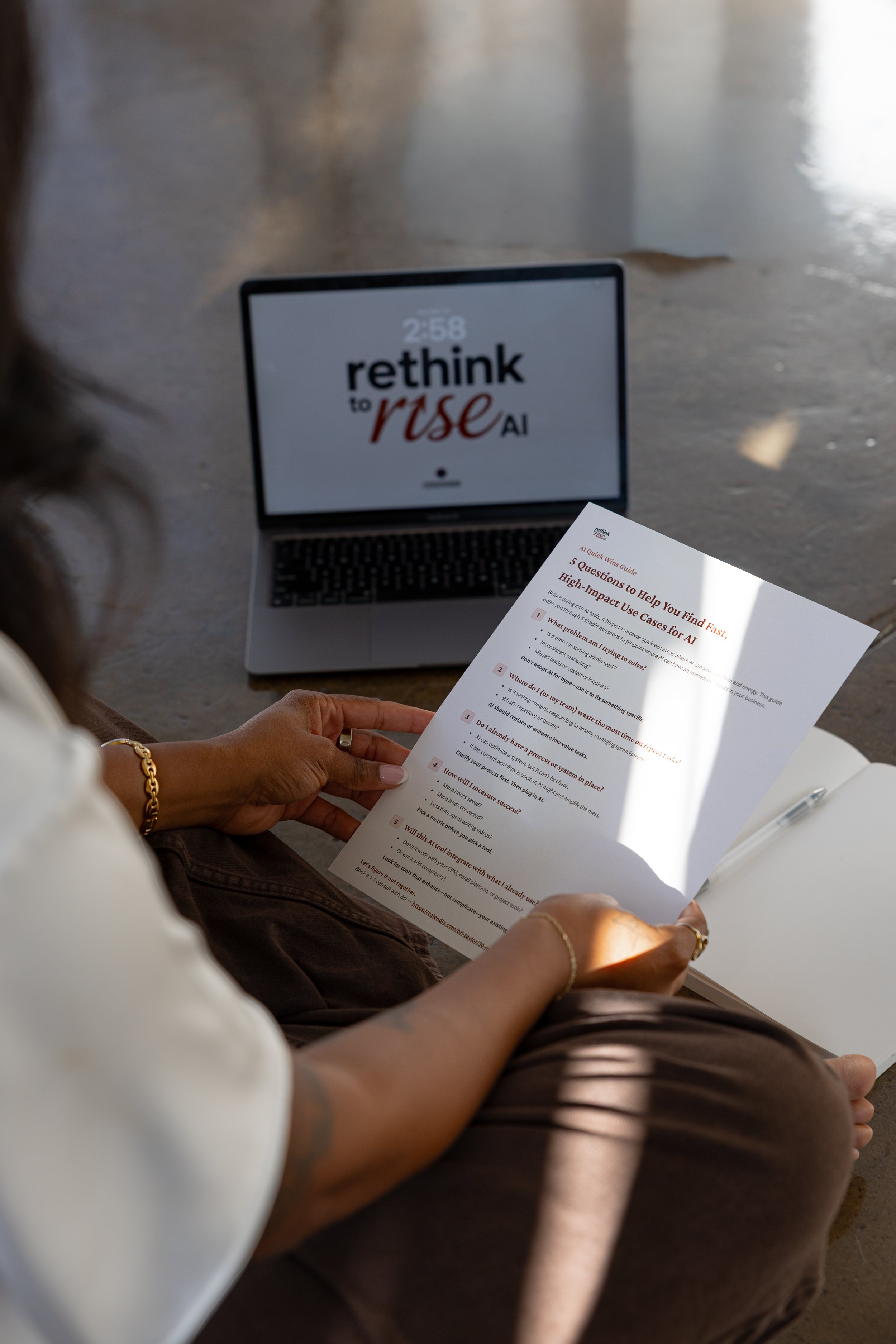 Person sitting on the floor reading a printed guide titled '5 Questions to Help You Find Fast, High-Impact Use Cases for AI' with a laptop in the background displaying 'rethink to rise AI' on the screen.