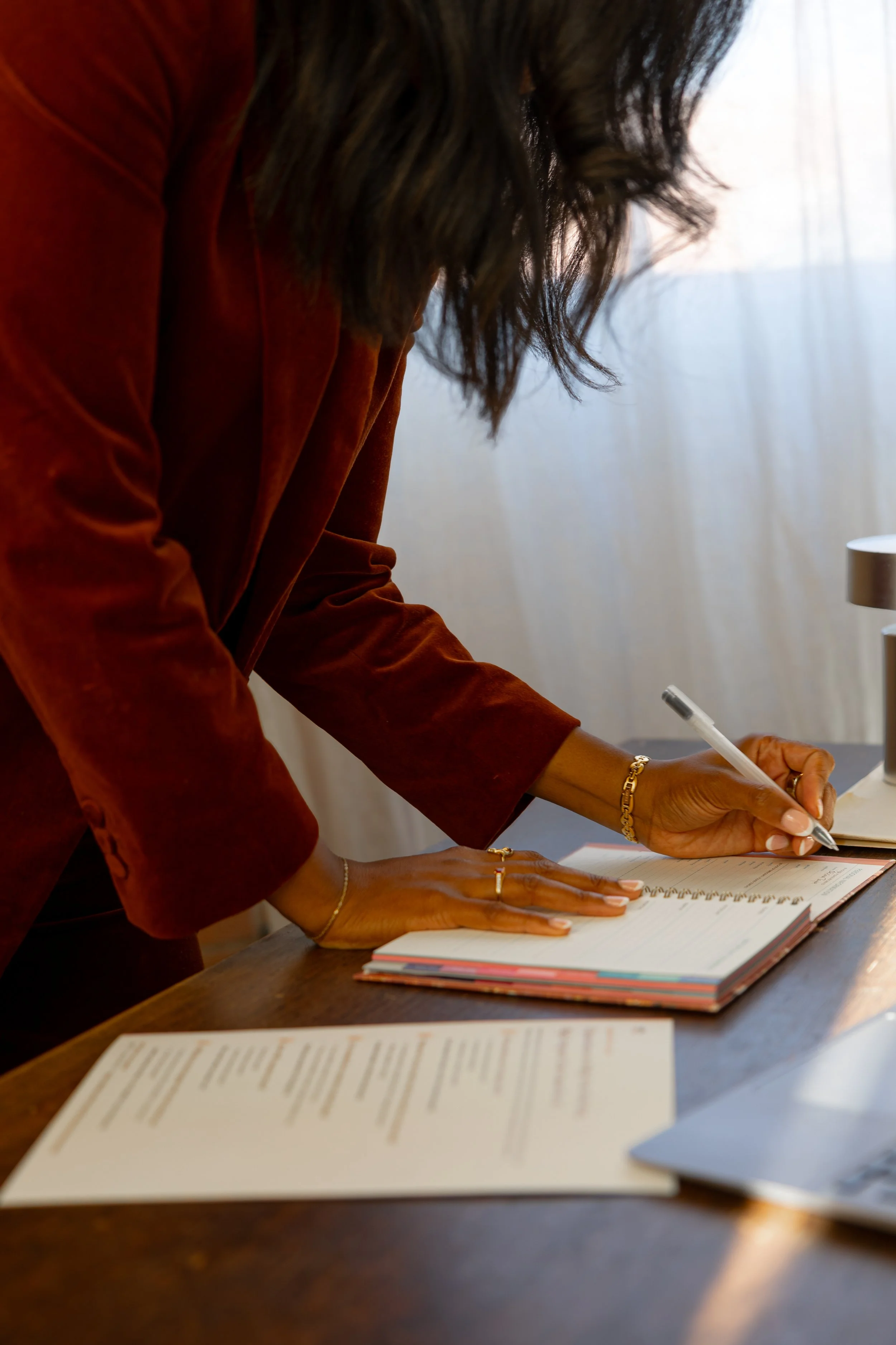 A woman in an orange blazer is writing in a planner on a wooden desk, with papers, a laptop, and a lamp nearby.