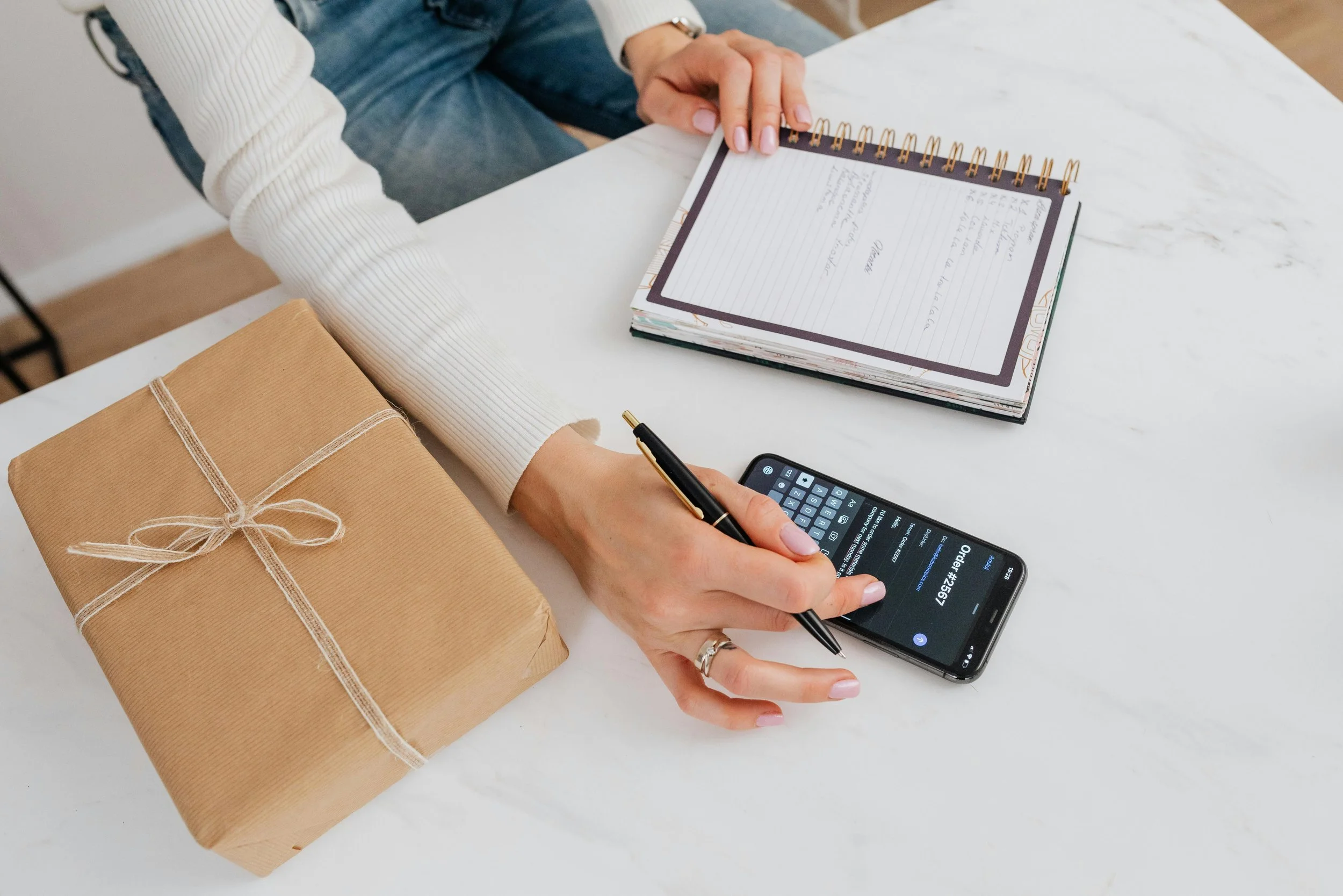 Person using a smartphone to check an order number, with a wrapped gift on a white table, an open planner, and a gold pen in hand. Rethink to Rise AI