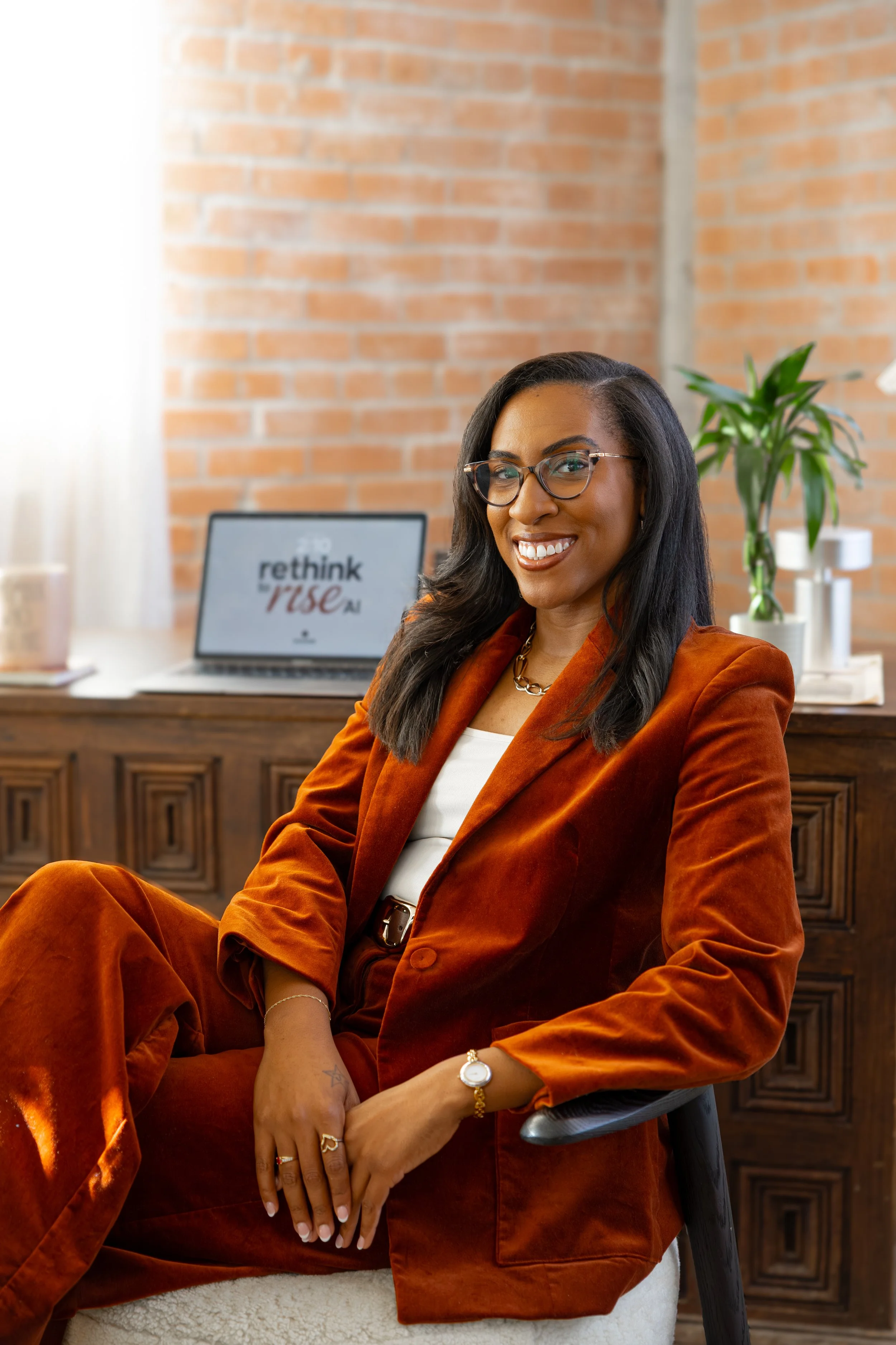 A confident woman with dark hair, glasses, and a warm smile sitting in an office with a brick wall background. She is wearing an orange velvet suit and accessorized with jewelry. A laptop displaying 'rethink & rise Ai' is on a wooden desk behind her, along with a potted plant.