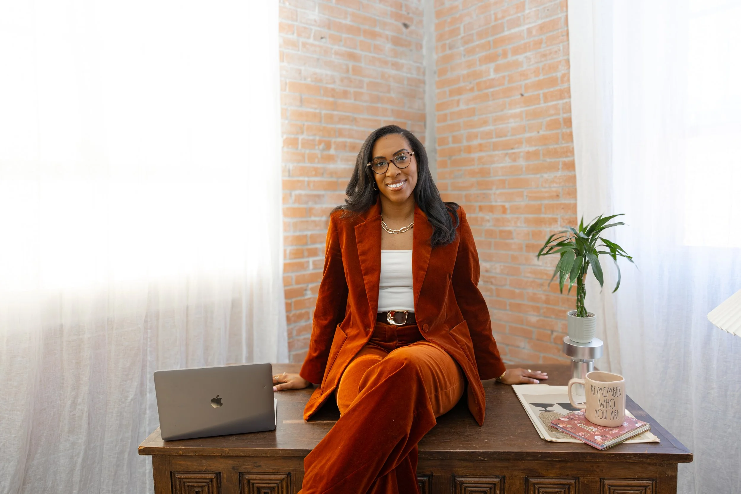 A woman in a rust-colored suit and white top sitting on a wooden desk in a room with brick walls and white curtains, smiling at the camera.