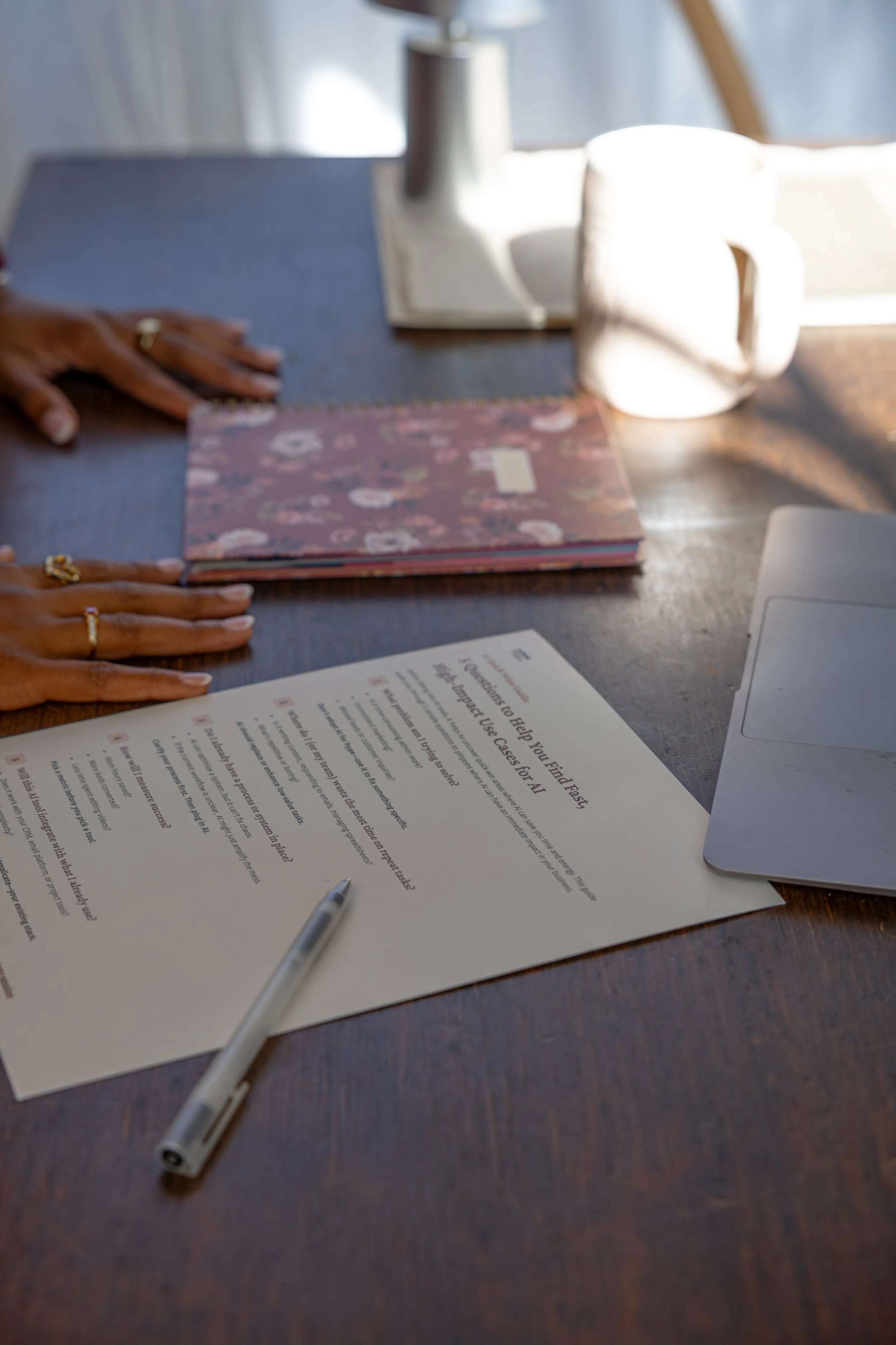 A wooden desk with a document, a pen, a laptop, a floral notebook, a coffee mug, a book, and a person's hands resting on the desk.