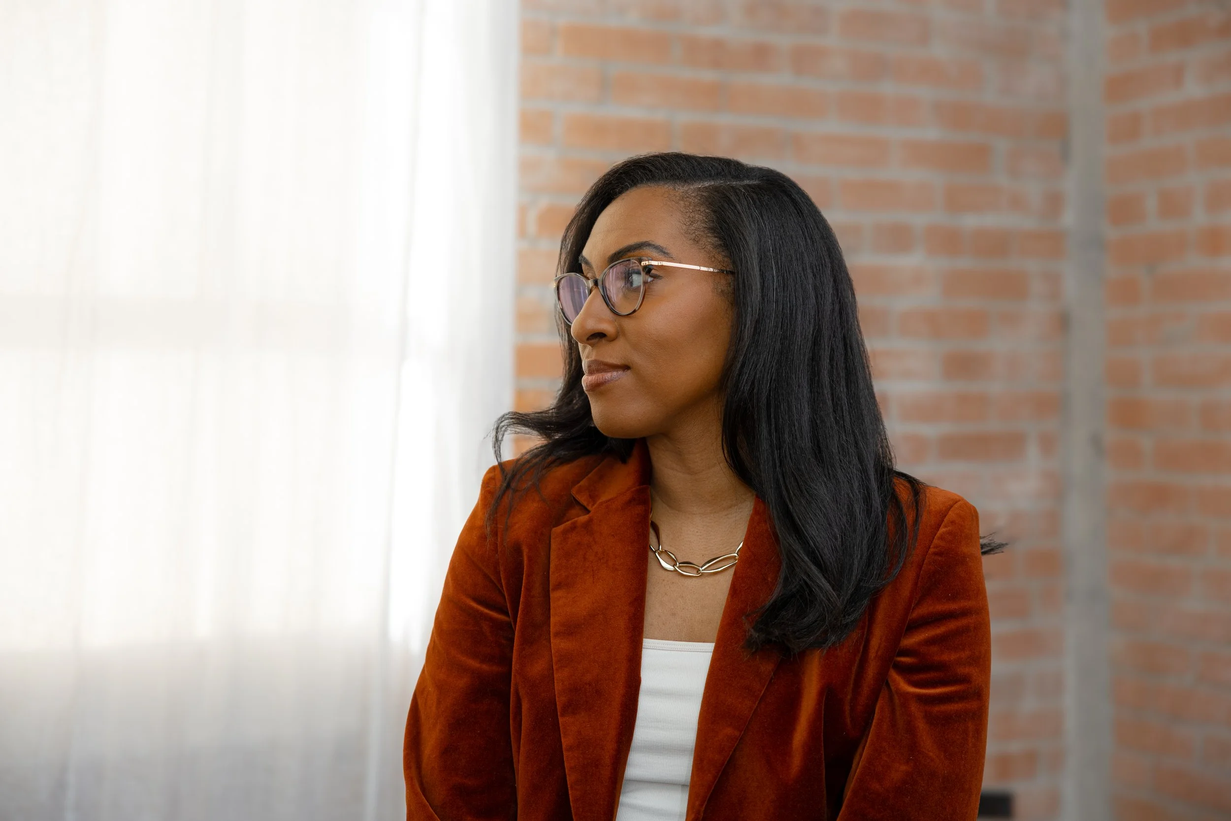 A woman with long black hair, glasses, wearing a rust-colored blazer and white top, looking to the side with a neutral expression, in front of a brick wall and sheer white curtains.