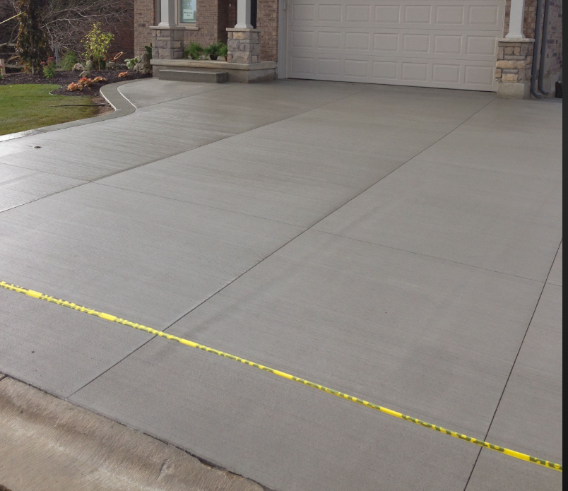 Newly poured concrete driveway in front of a house with a garage, bordered by a yellow measurement tape, and garden with plants and mulch on the side.