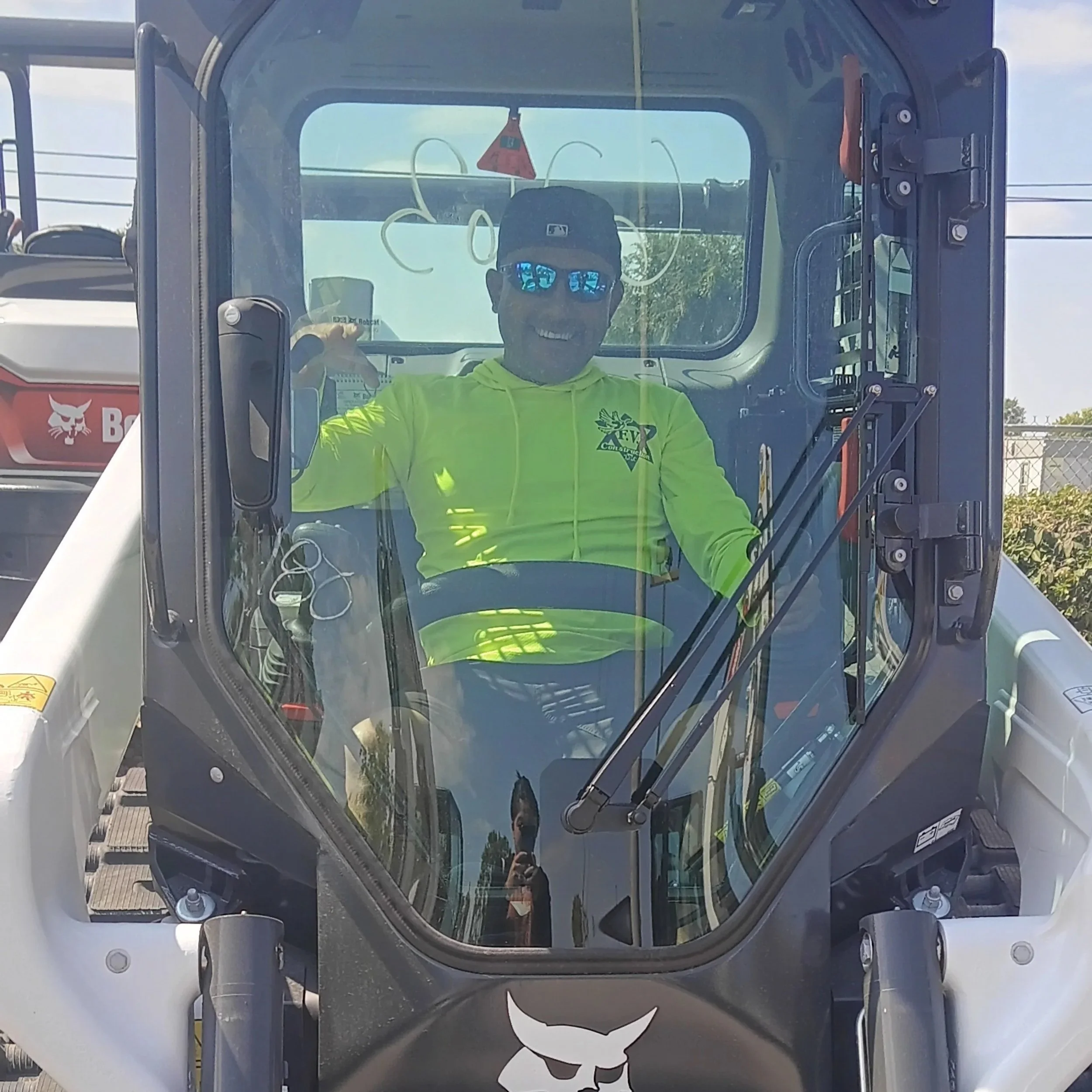 A smiling man wearing a yellow hoodie, black cap, and sunglasses sitting inside a construction vehicle, with a reflecty view of the photographer taking the photo.