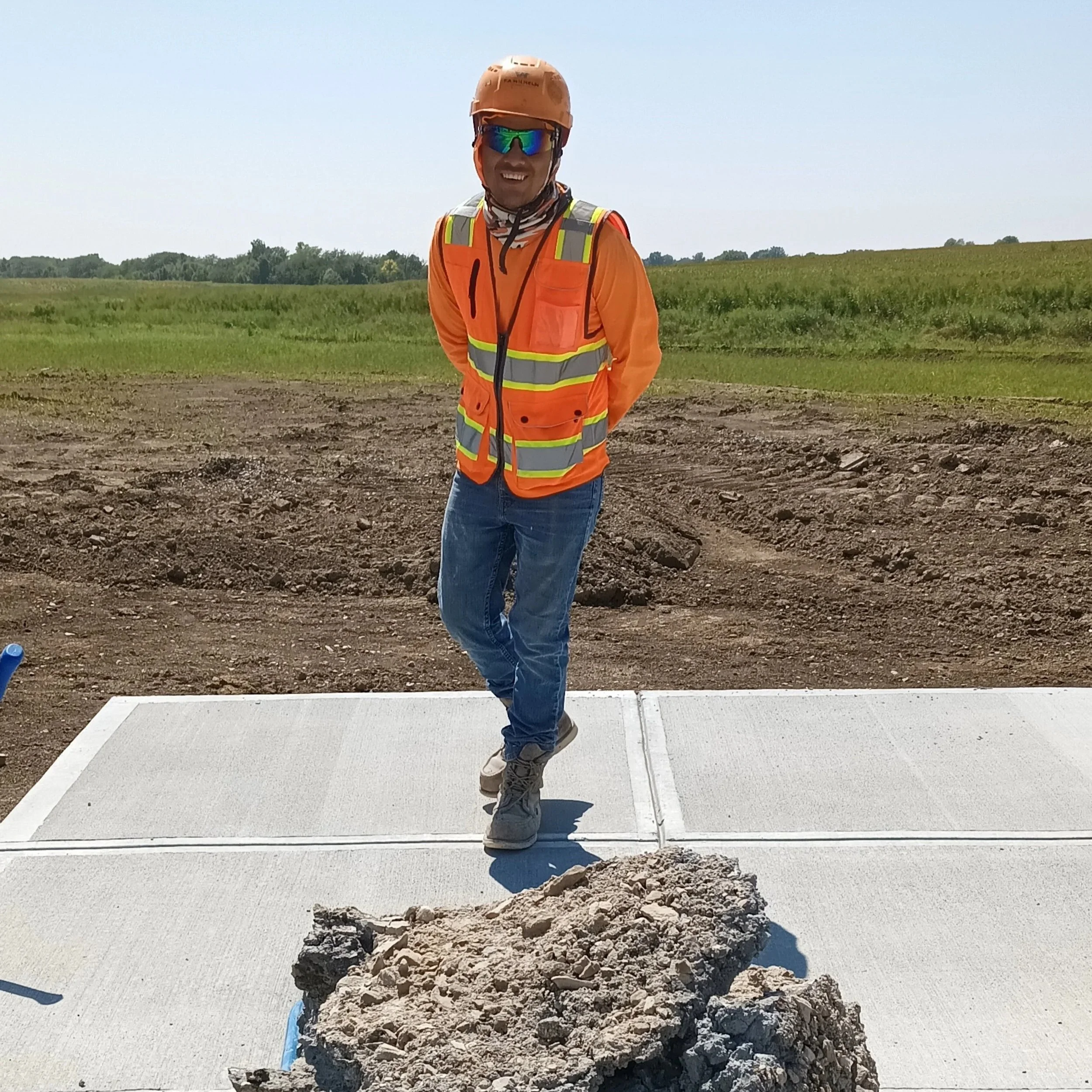 A man in construction safety gear, including an orange hard hat, reflective safety vest, and sunglasses, standing outdoors on a concrete slab with a large rock in front of him, with green fields in the background.