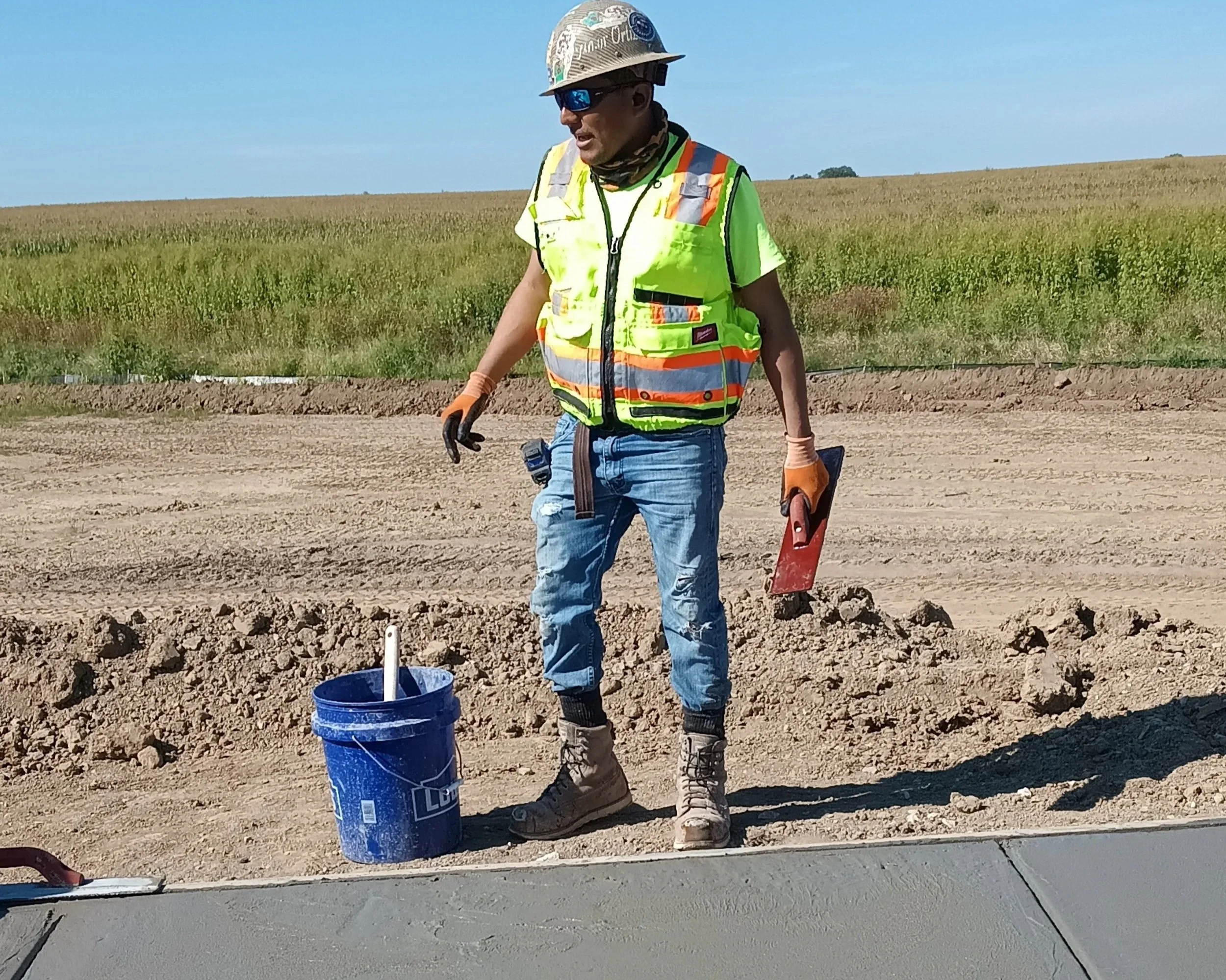 Construction worker wearing a yellow safety vest, hard hat, sunglasses, and work boots, working outdoors on a construction site with dirt and a concrete surface, holding a trowel, with a blue bucket nearby.