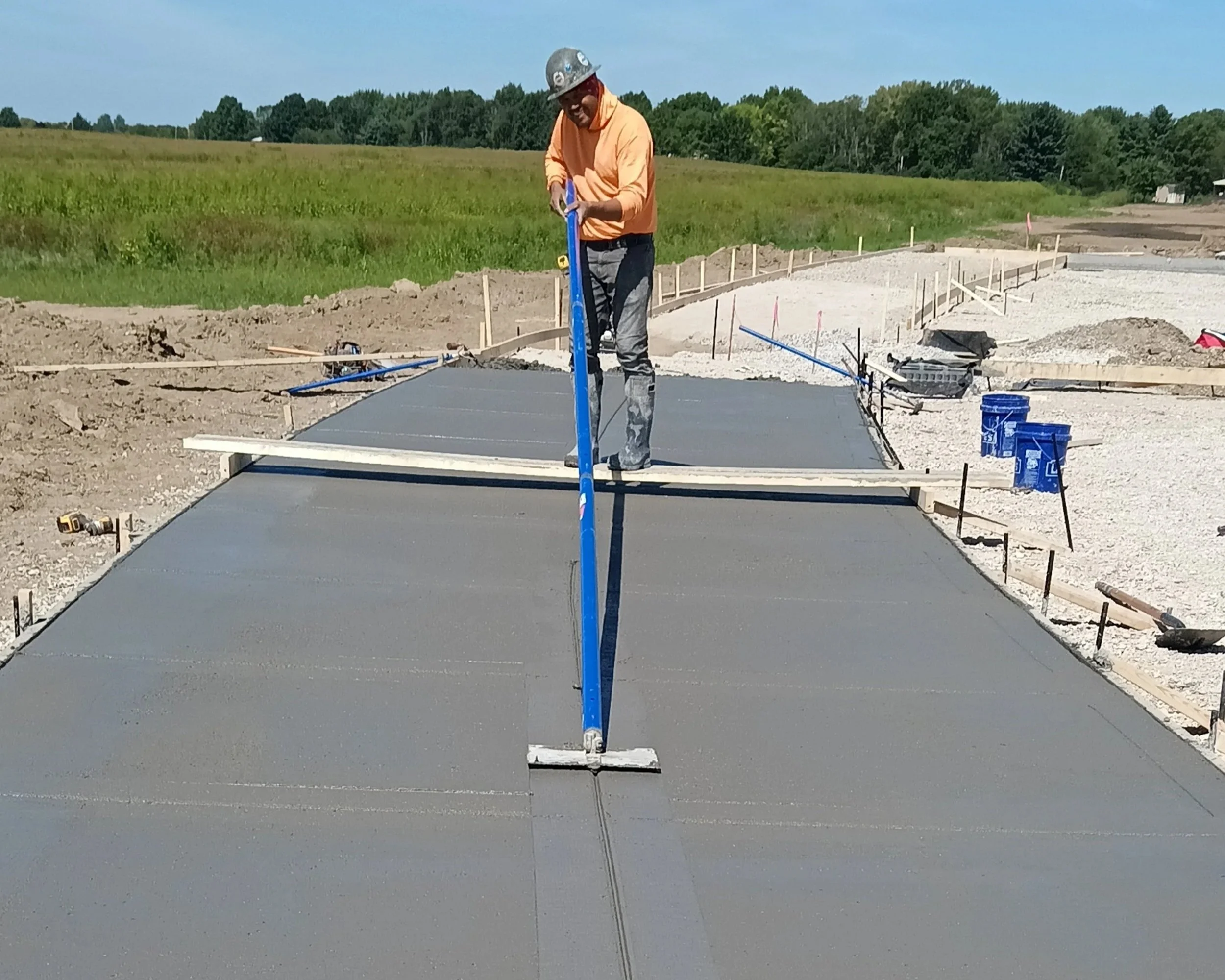 A construction worker in an orange shirt and gray pants, wearing a safety helmet, smooths wet concrete with a long leveling tool on a new driveway during construction.