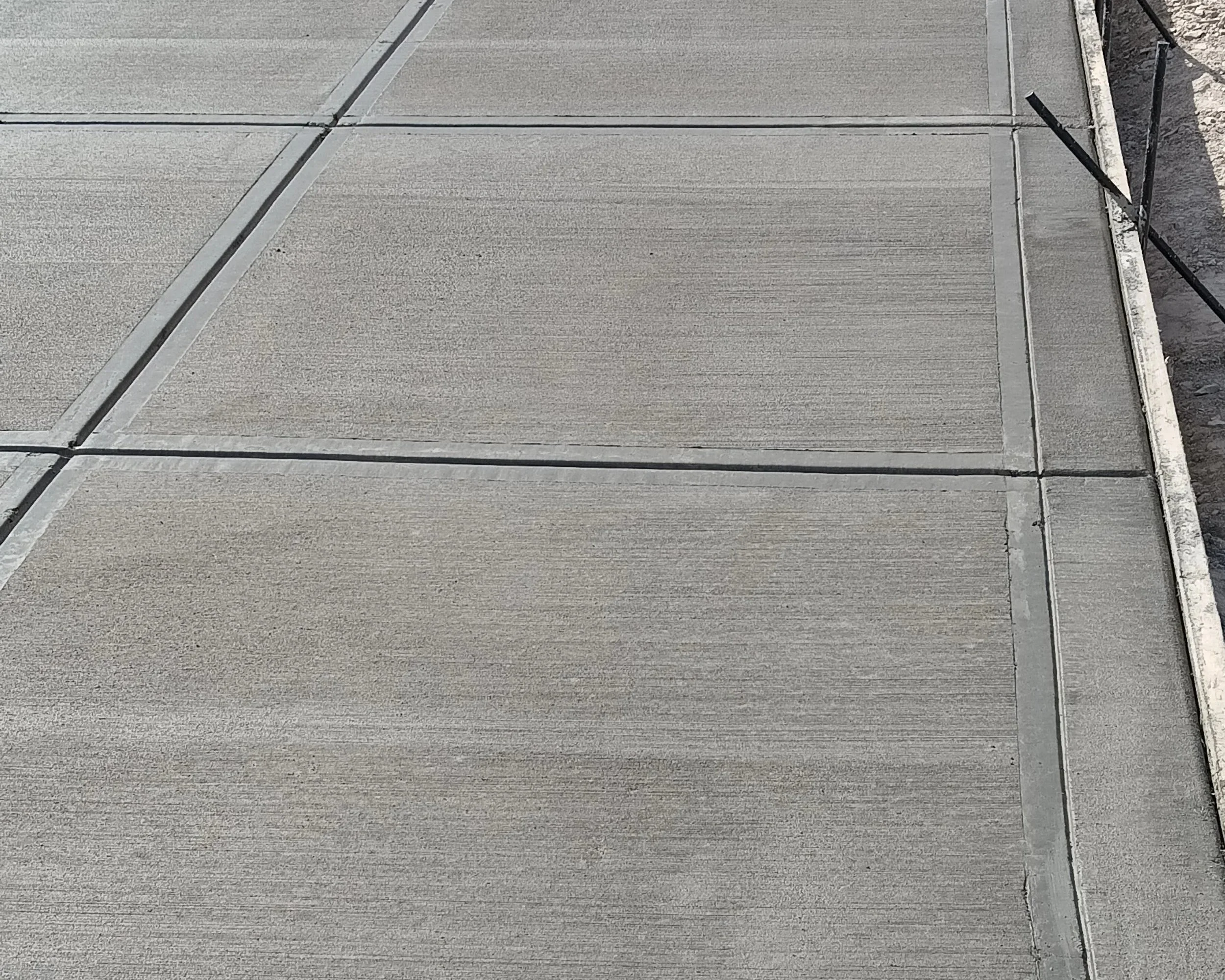 Newly poured concrete sidewalk with expansion joints and formwork on the right edge, outdoor setting.