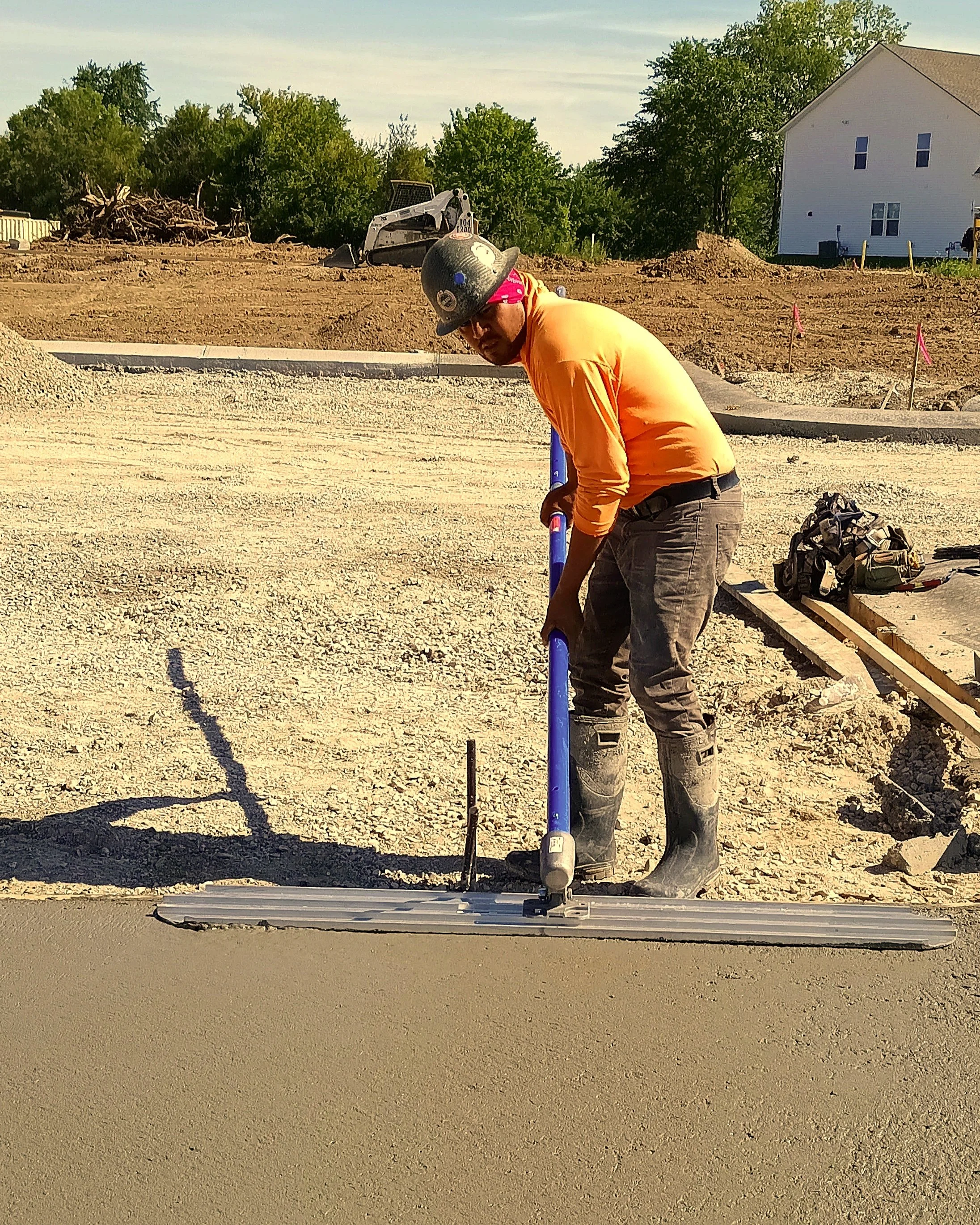 Construction worker using a hand tamper to level freshly poured concrete on a building site, wearing a helmet, orange shirt, and boots.