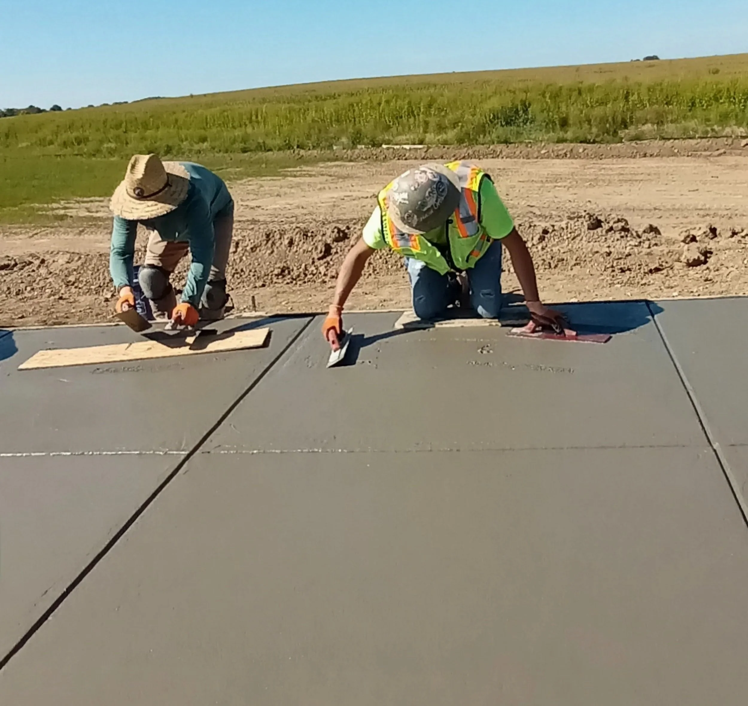 Two construction workers smoothing and finishing a concrete sidewalk in an open outdoor area with grassy fields in the background.