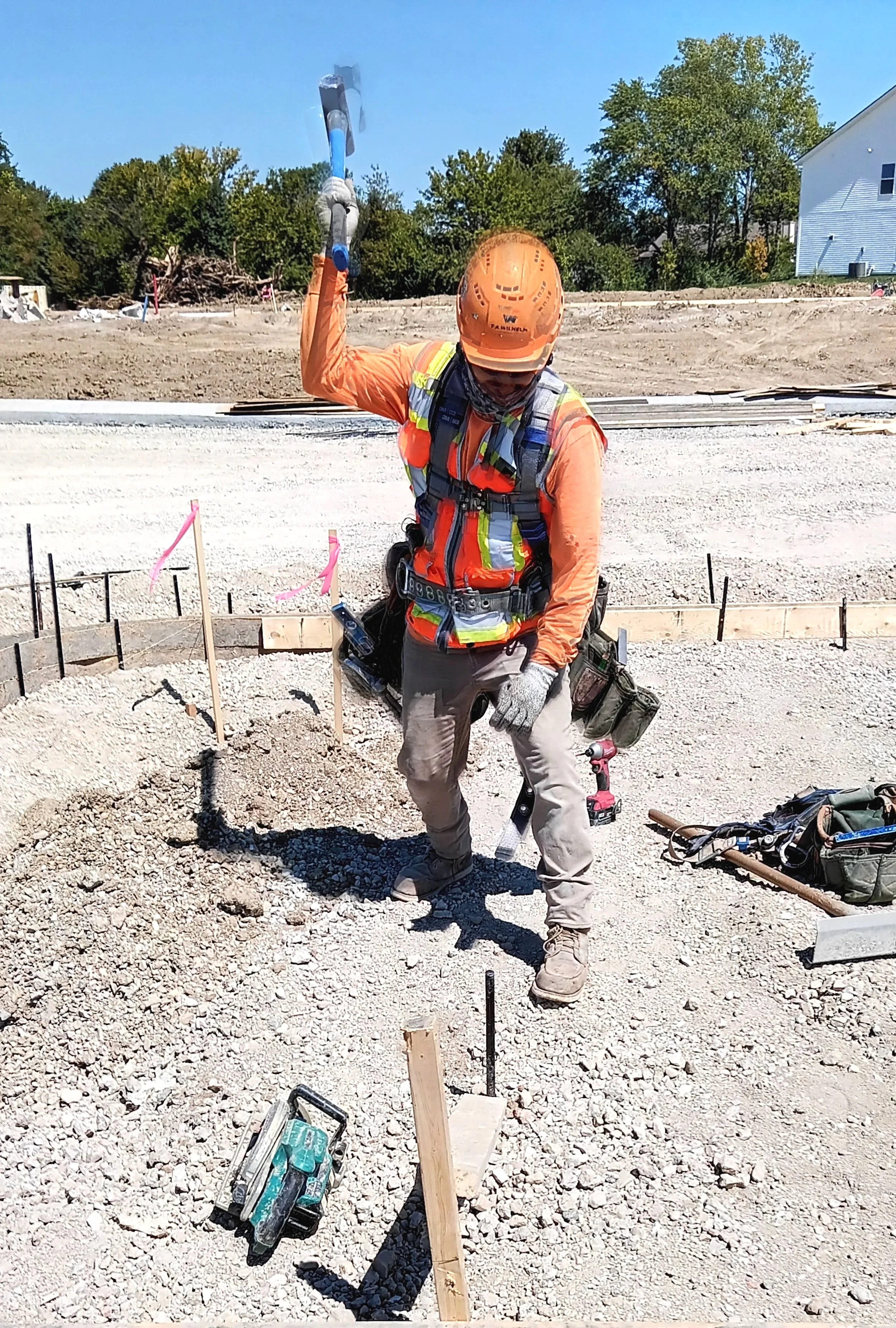 Construction worker in safety gear swinging a sledgehammer at a construction site with dirt and gravel ground and trees and a white house in the background.
