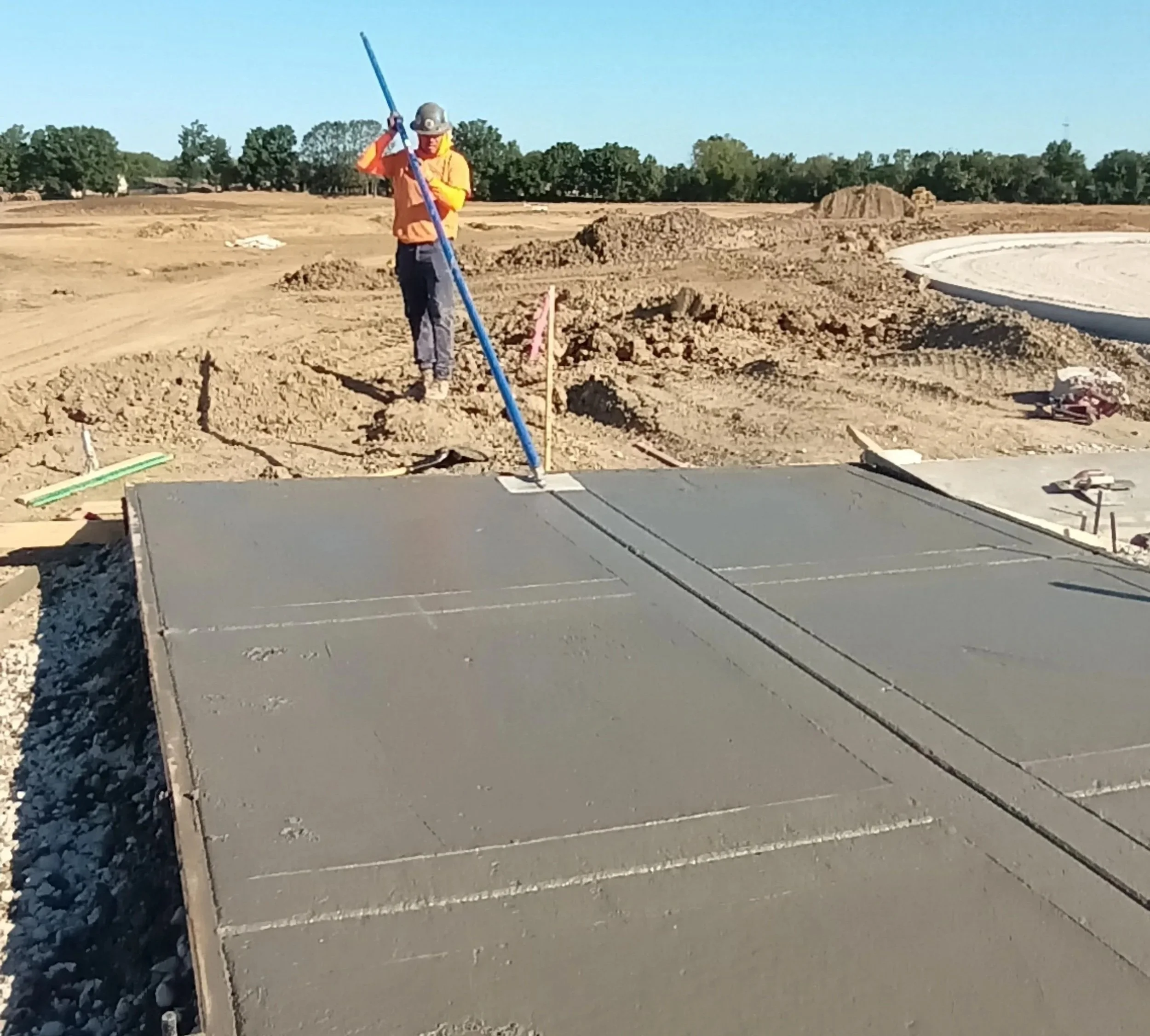 A construction worker smoothing wet concrete on a large slab with a long pole at a construction site on a sunny day.