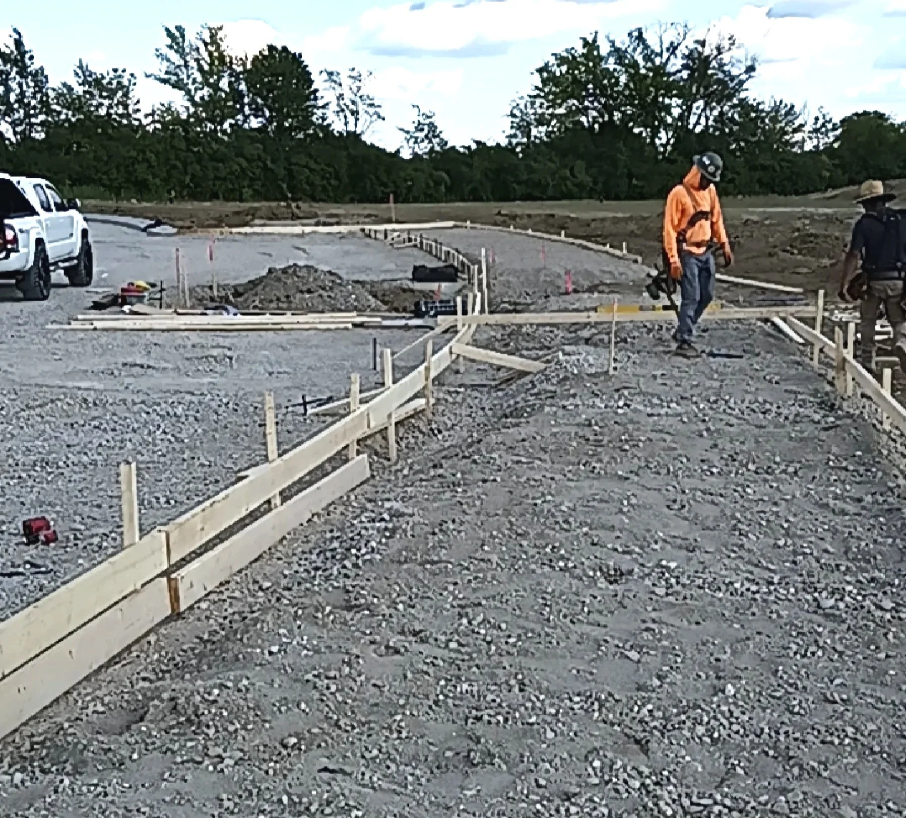 Construction workers walking on an unfinished road with wooden barriers, construction equipment, and a parked vehicle in the background.