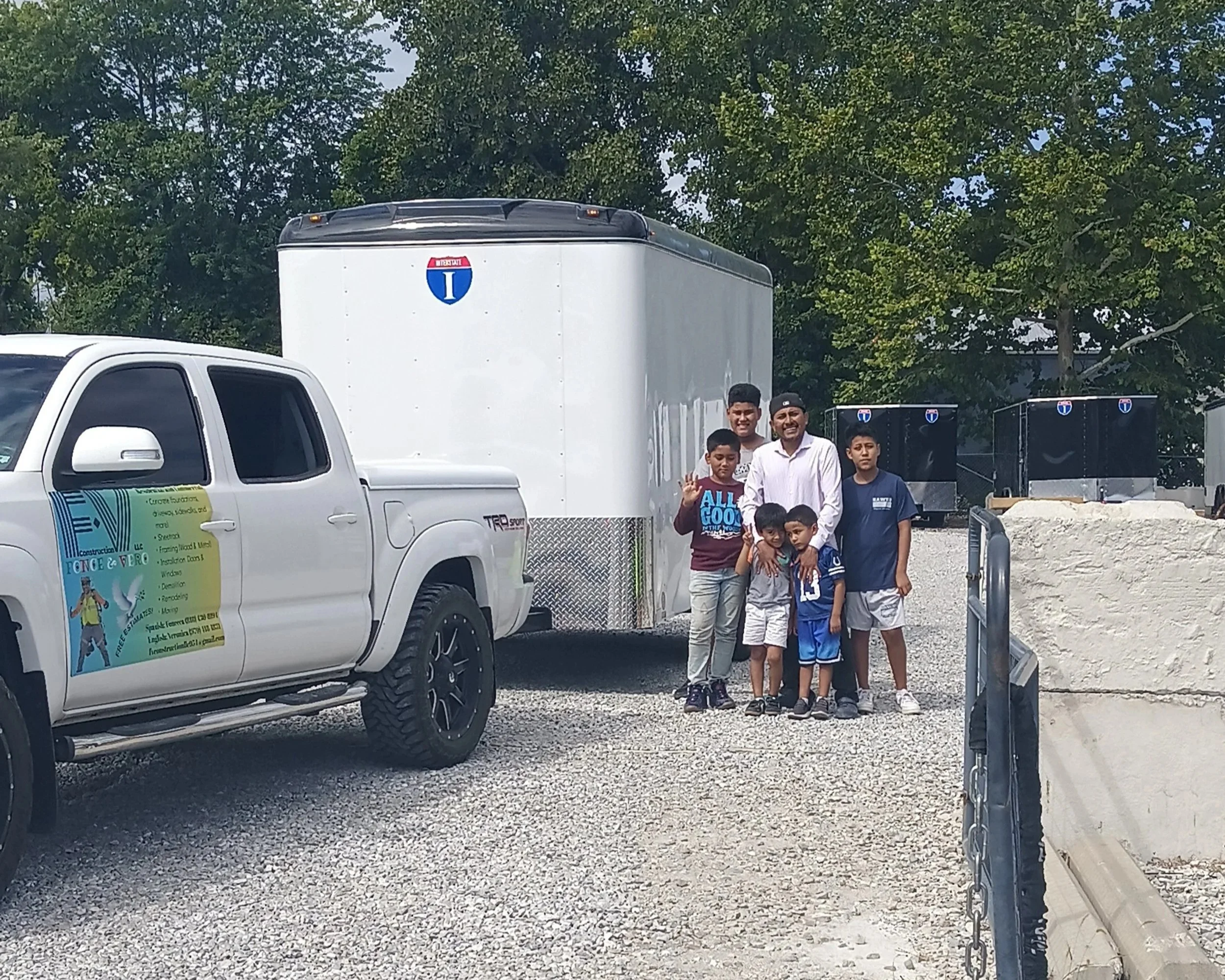 Group of five boys and one adult man standing outdoors on a gravel lot next to a white pickup truck with a construction advertisement and a large white trailer with a blue and red logo, surrounded by trees.