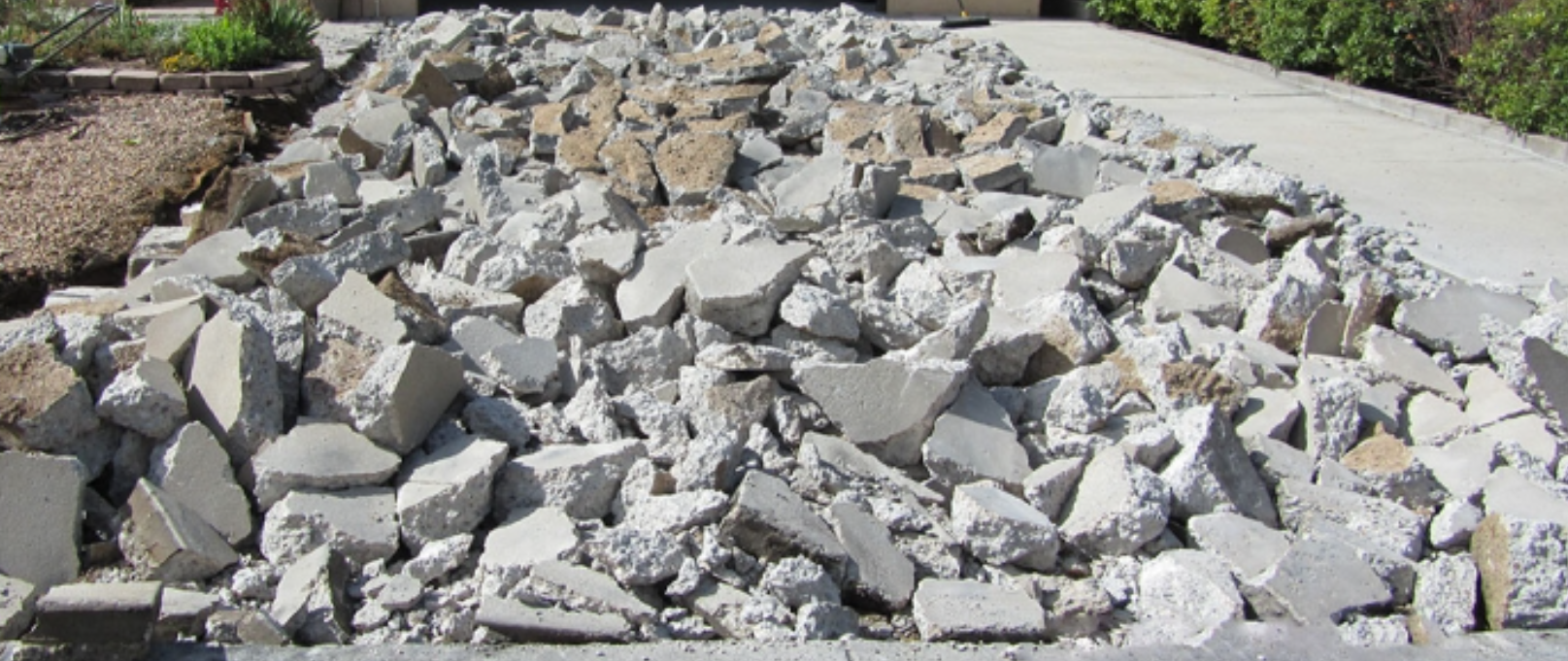 A pile of broken concrete and rocks in front of a sidewalk with plants on the side.