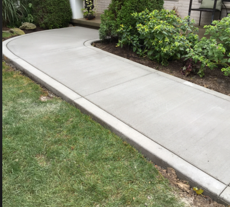 Concrete sidewalk curving past a grassy lawn and bushes near a house with a porch and outdoor furniture.