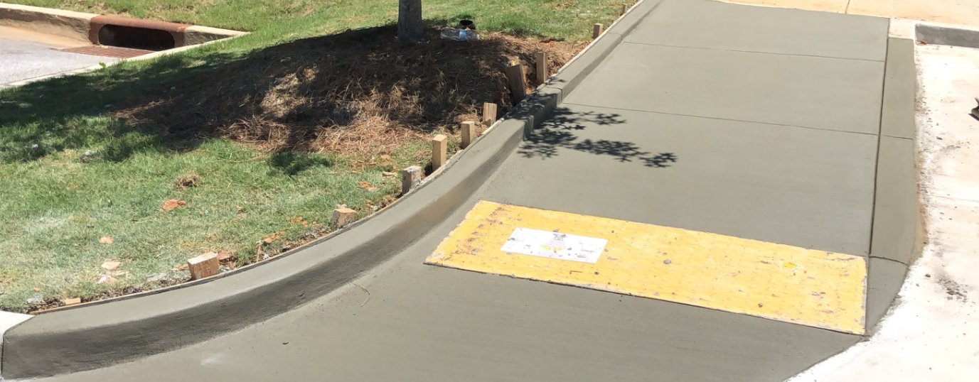 Newly constructed concrete sidewalk with a yellow tactile warning strip for visually impaired pedestrians, nearby grassy area, and shadow of a tree.
