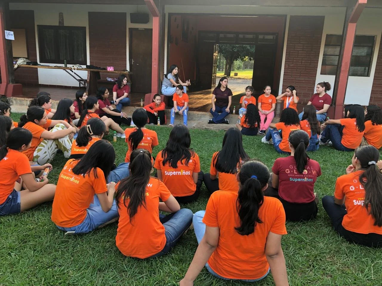 Group of girls sitting on grass in front of a building, participating in an outdoor event, some wearing orange shirts with the words 'Superninhos' printed on the back.