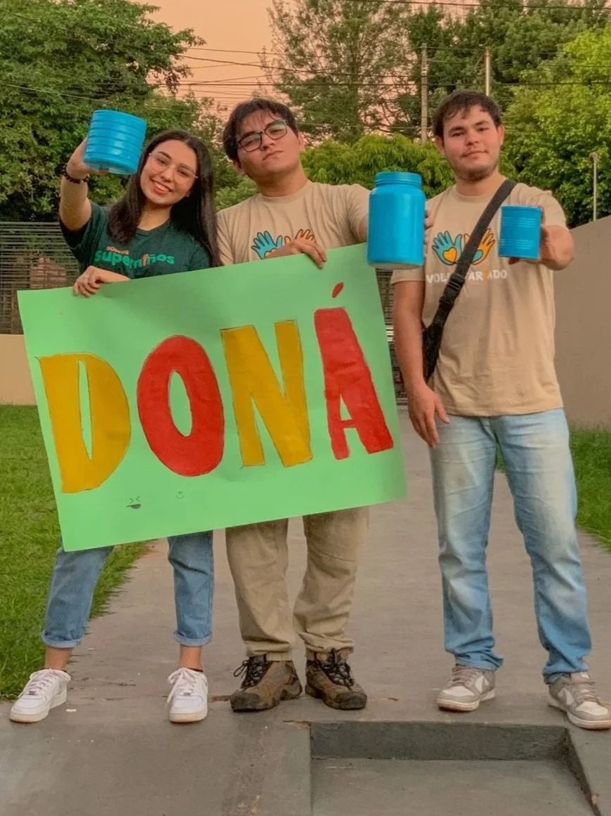 Three young people standing outdoors on a sidewalk, holding a green sign with the word 'DONA' in colorful letters. The person on the left is smiling and holding a blue container, the person in the middle has glasses and a serious expression, holding 