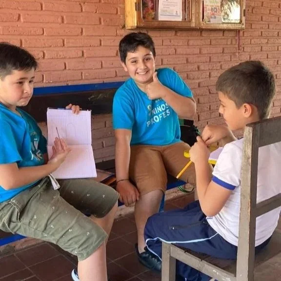Three boys sitting and chatting outdoors, one holding a notebook and pen, smiling