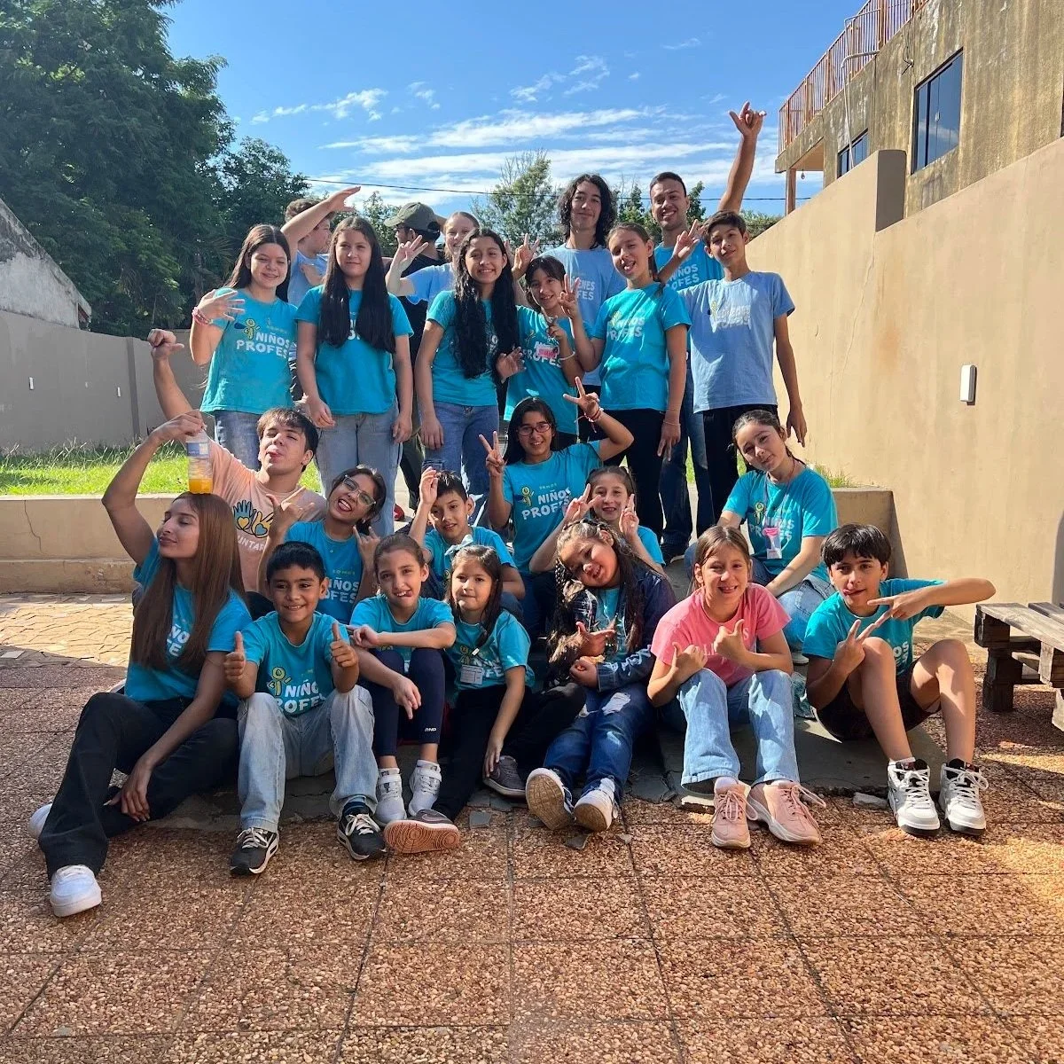Group of children and teenagers smiling and posing outdoors during daytime, some wearing matching blue t-shirts.