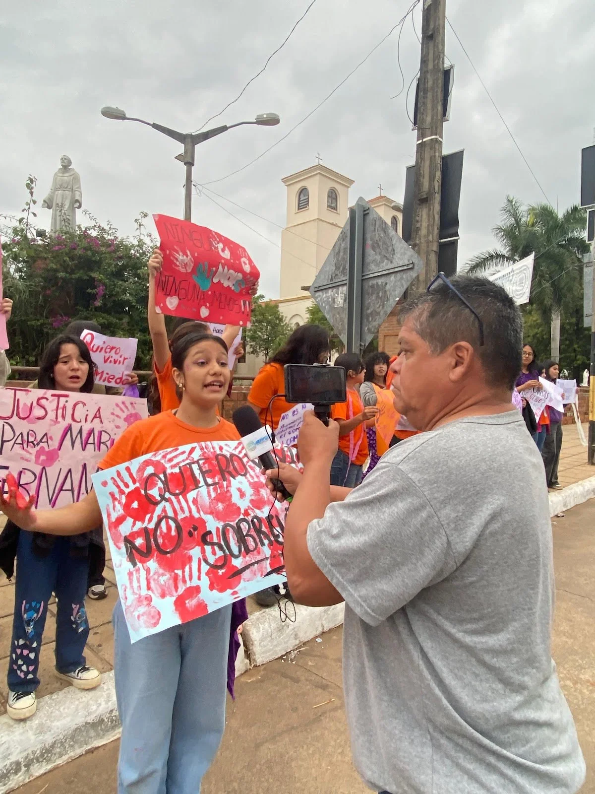 A young girl wearing an orange shirt holding a protest sign and speaking to a man with a video camera during a protest. Several other protesters behind her hold signs and banners, and there is a church with a bell tower in the background.